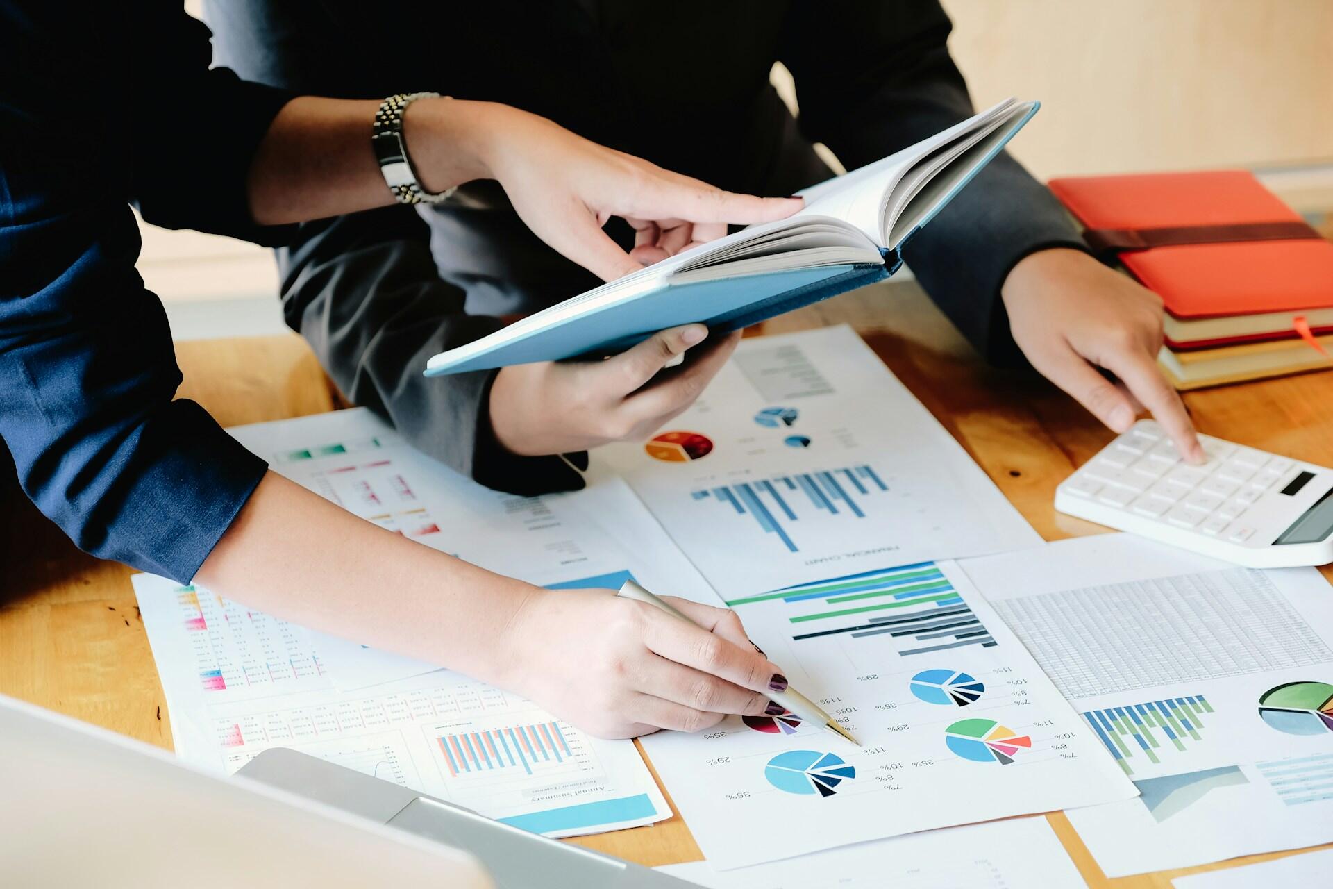 Dos profesionales analizan tablas y gráficos en documentos mientras uno sostiene un libro abierto, colaborando sobre una mesa de madera.