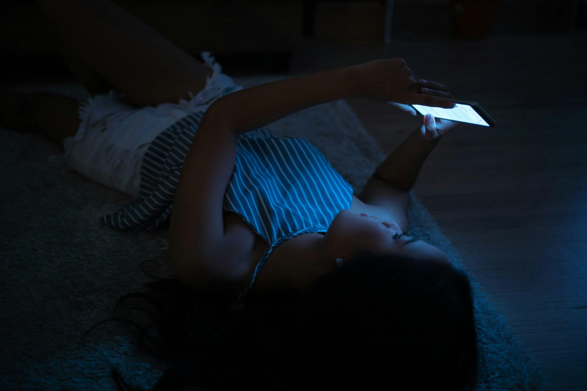Mujer acostada en el suelo en la oscuridad usando un celular. La luz azul de la pantalla ilumina su rostro. Lleva blusa de rayas y shorts blancos. Escena nocturna que refleja uso de tecnología antes de dormir.