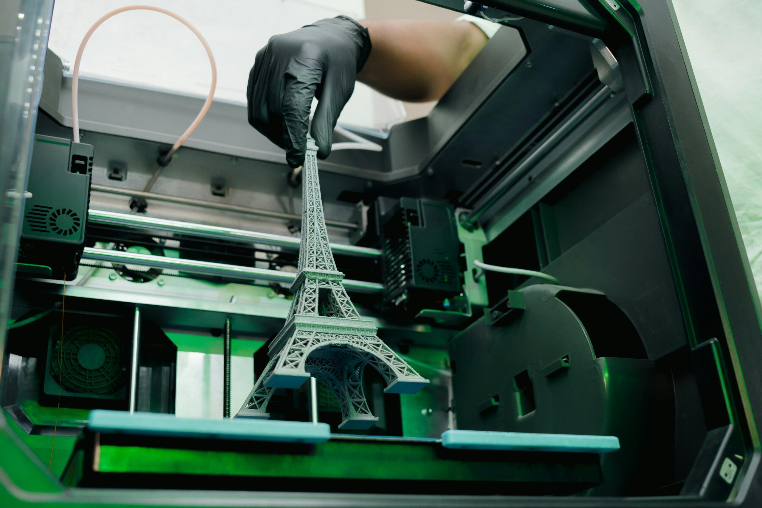 Una mano con un guante negro saca un modelo gris de la Torre Eiffel impreso en 3D de una impresora 3D con el interior iluminado en verde.