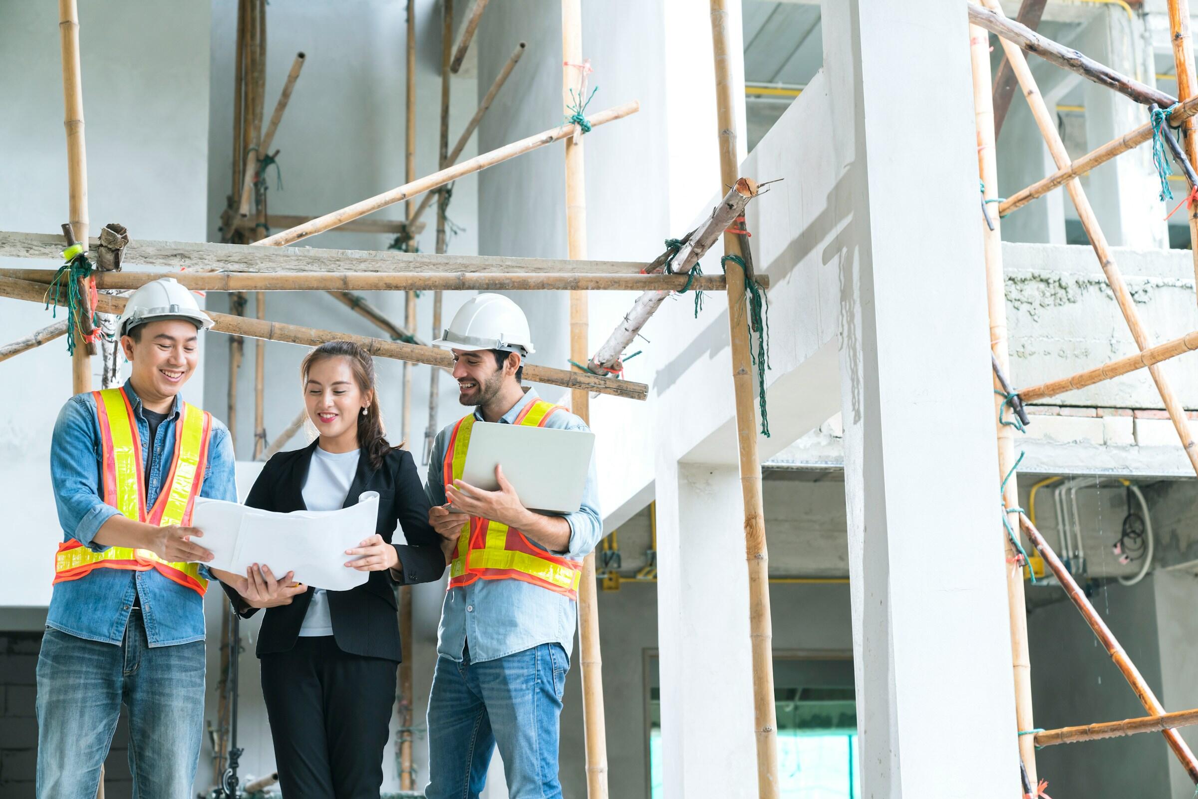 Tres profesionales de la construcción, equipados con material de seguridad, discuten los planos entre los andamios de una obra luminosa y sin terminar.