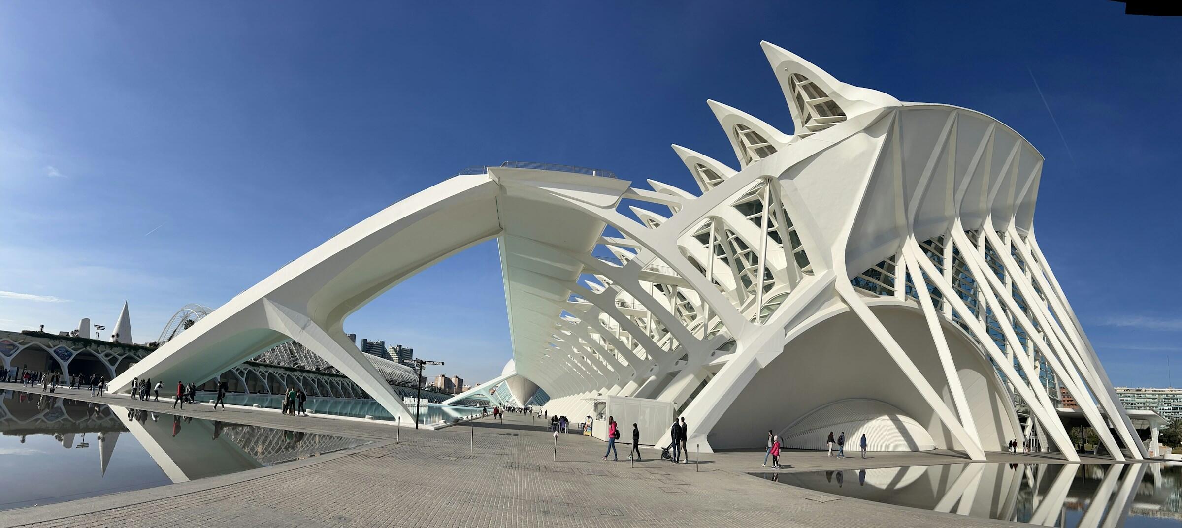 Una llamativa estructura arquitectónica de moderna con formas blancas y curvas de la ciudad de las artes y las ciencias de valencia, recortada contra un cielo azul claro, reflejada en una tranquila superficie de agua.