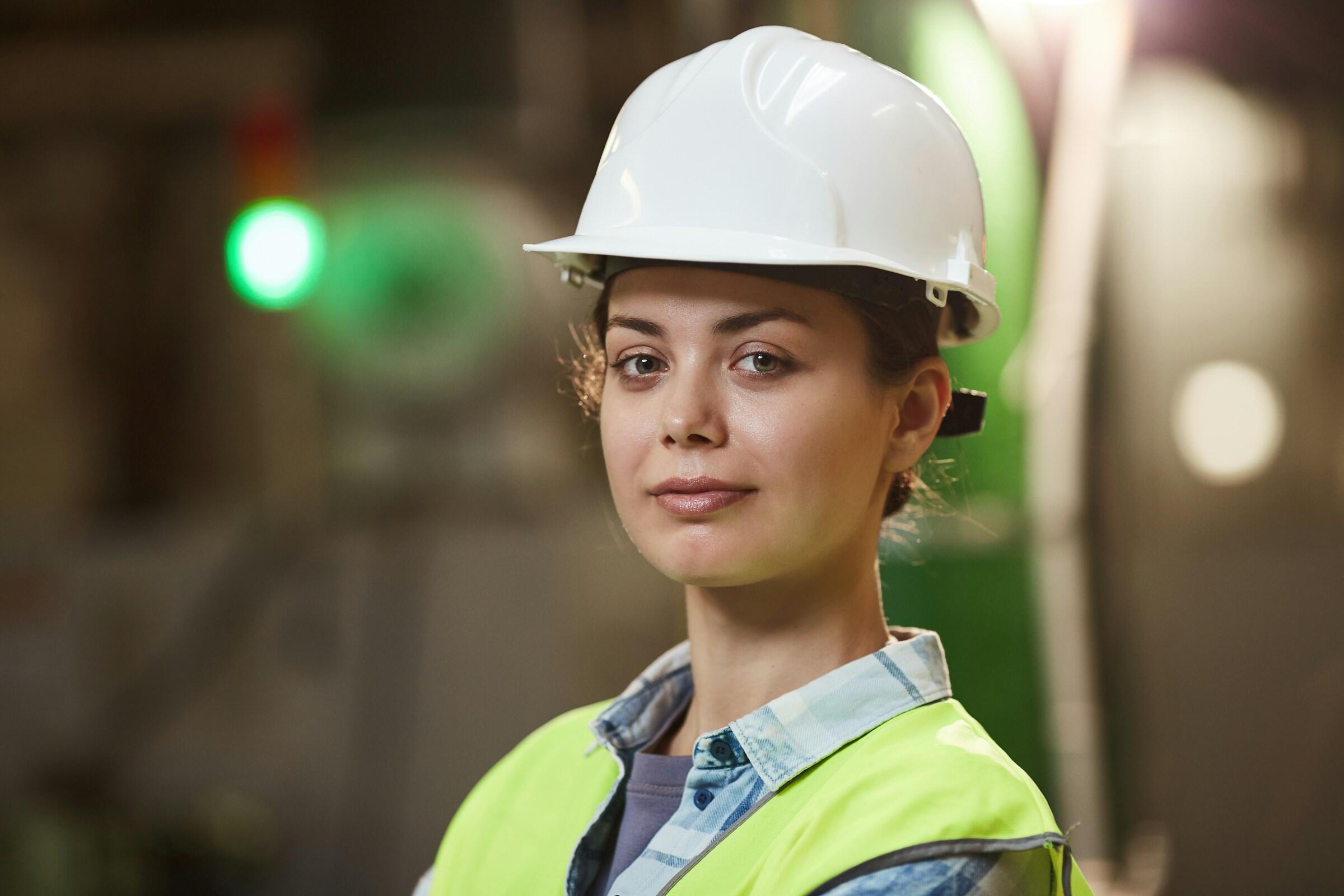 Una trabajadora de la construcción con un casco blanco y un chaleco de seguridad amarillo brillante posa con confianza en un entorno industrial bien iluminado.
