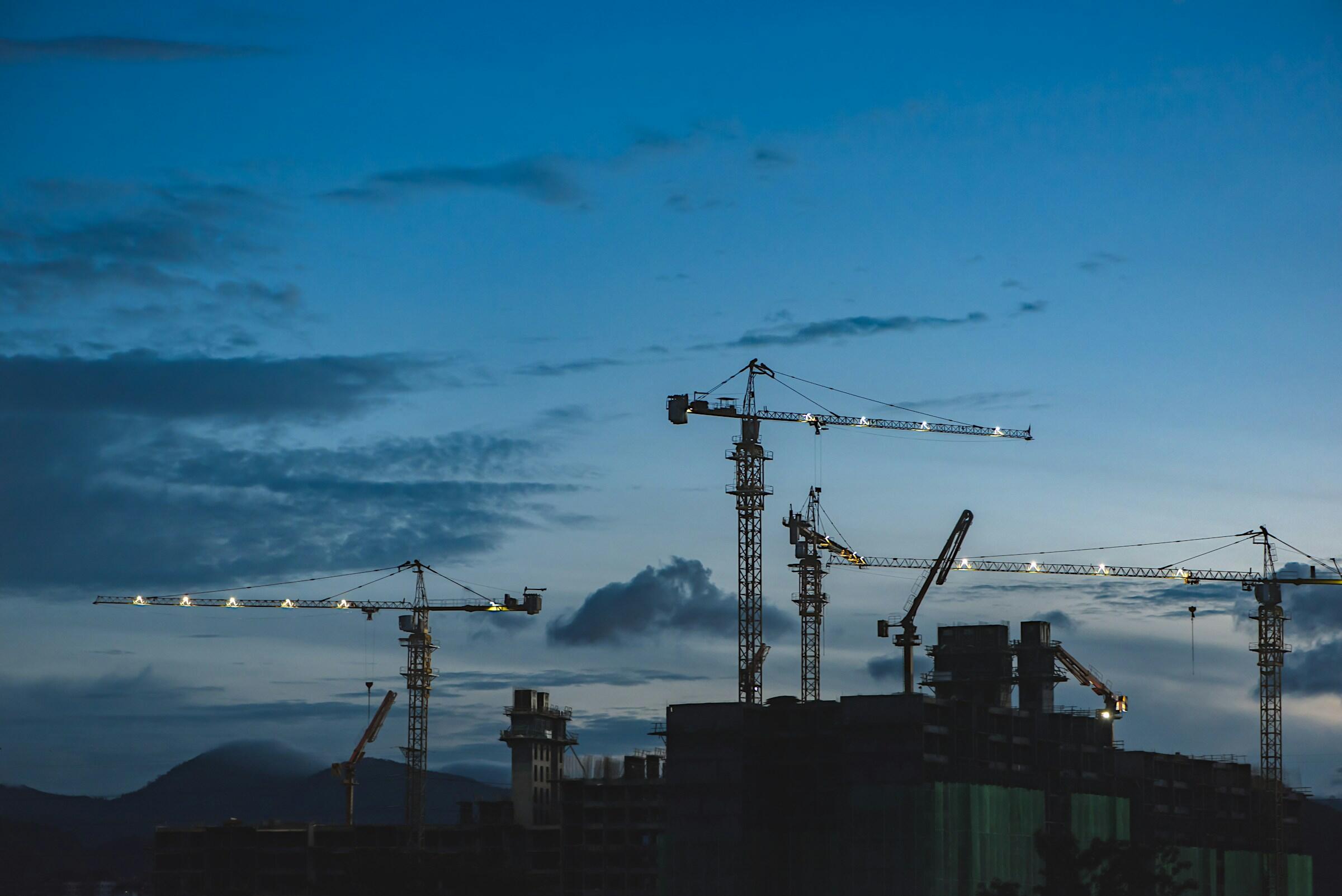 Grúas de construcción recortadas contra un cielo crepuscular, con nubes y montañas lejanas al fondo.