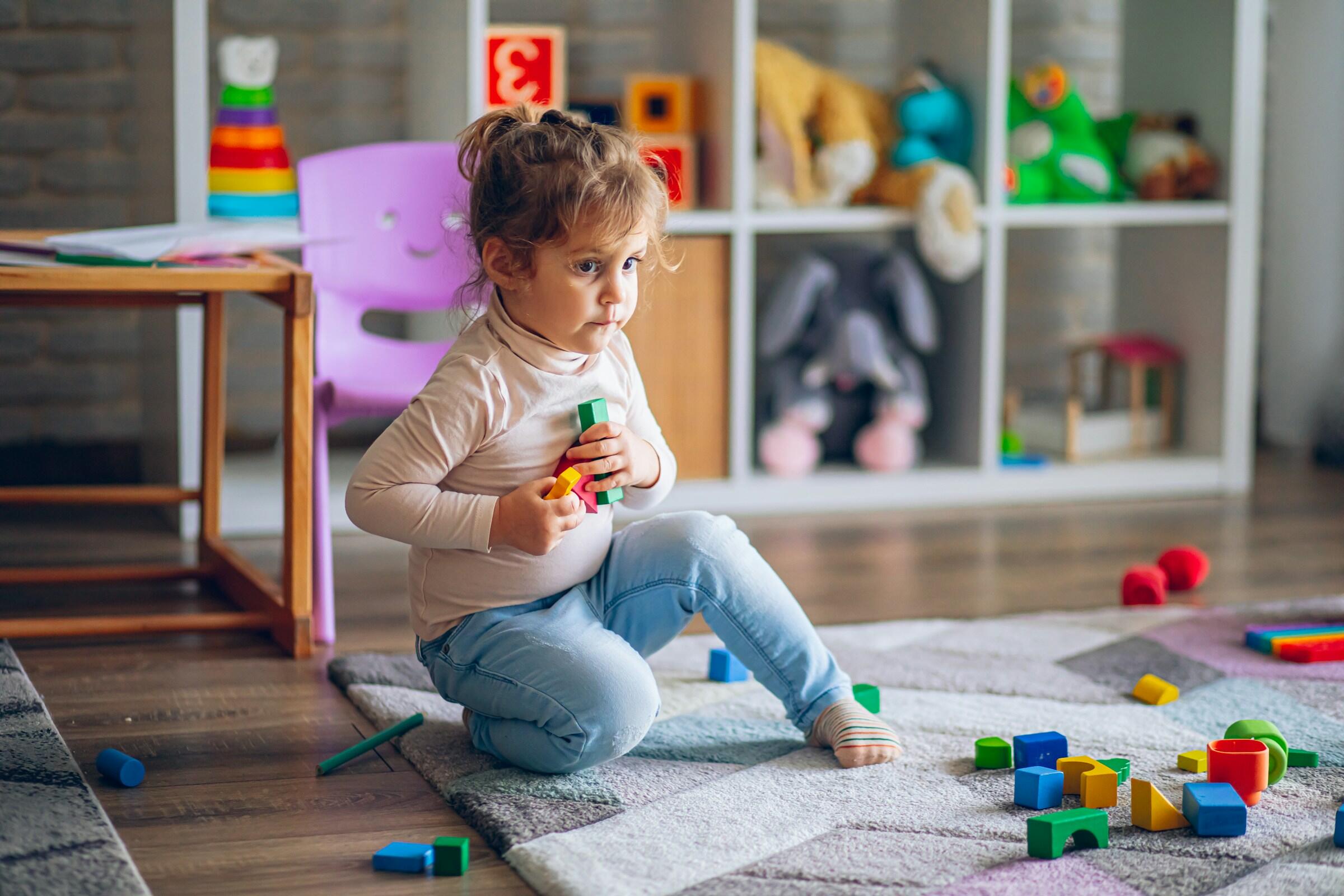 Un niño está sentado sobre una alfombra de colores jugando con bloques de construcción, rodeado de varios juguetes y una mesita en una luminosa sala de juegos.