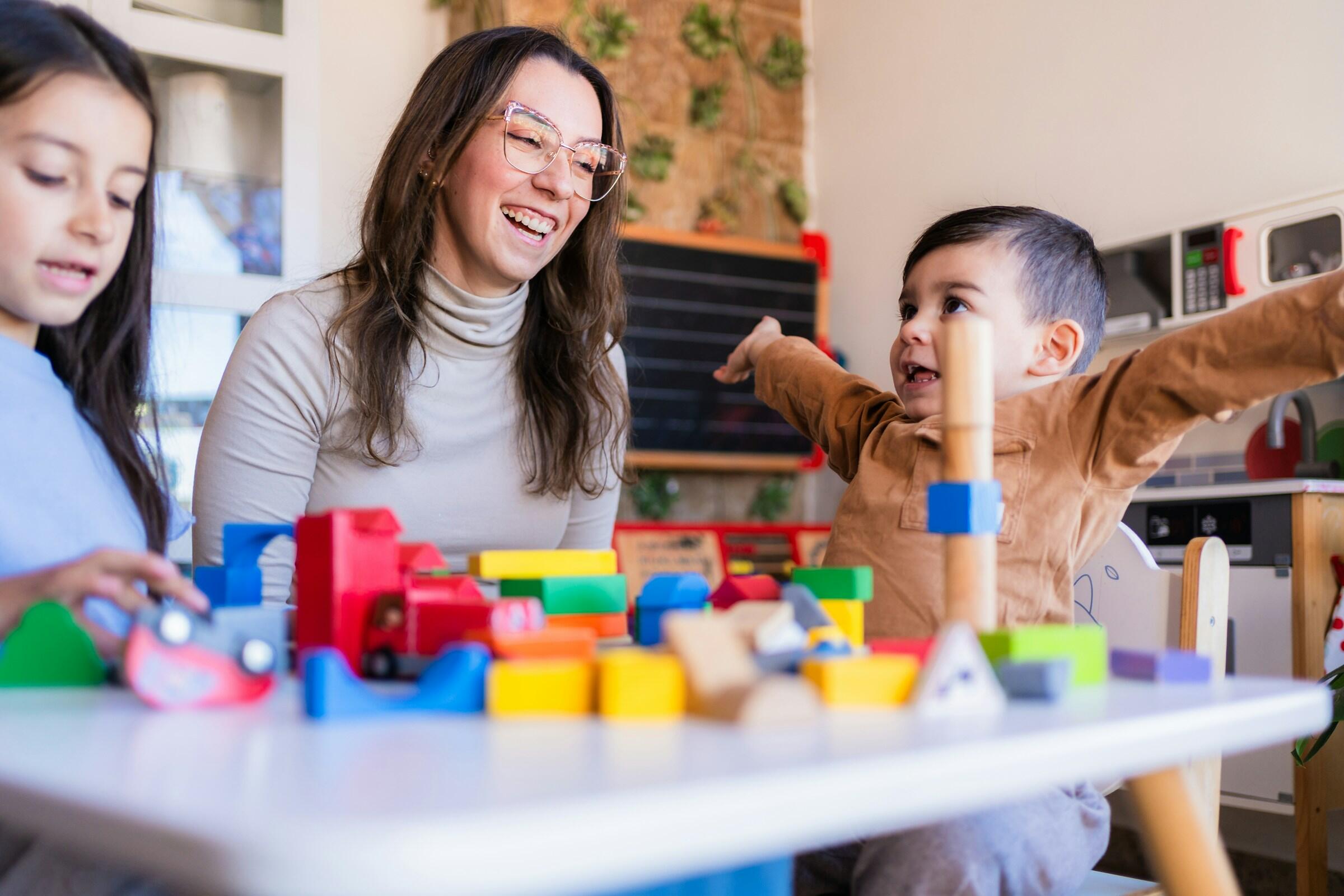 Una mesa llena de bloques de construcción y juguetes de colores, con dos niños jugando y una mujer observándolos a su lado.