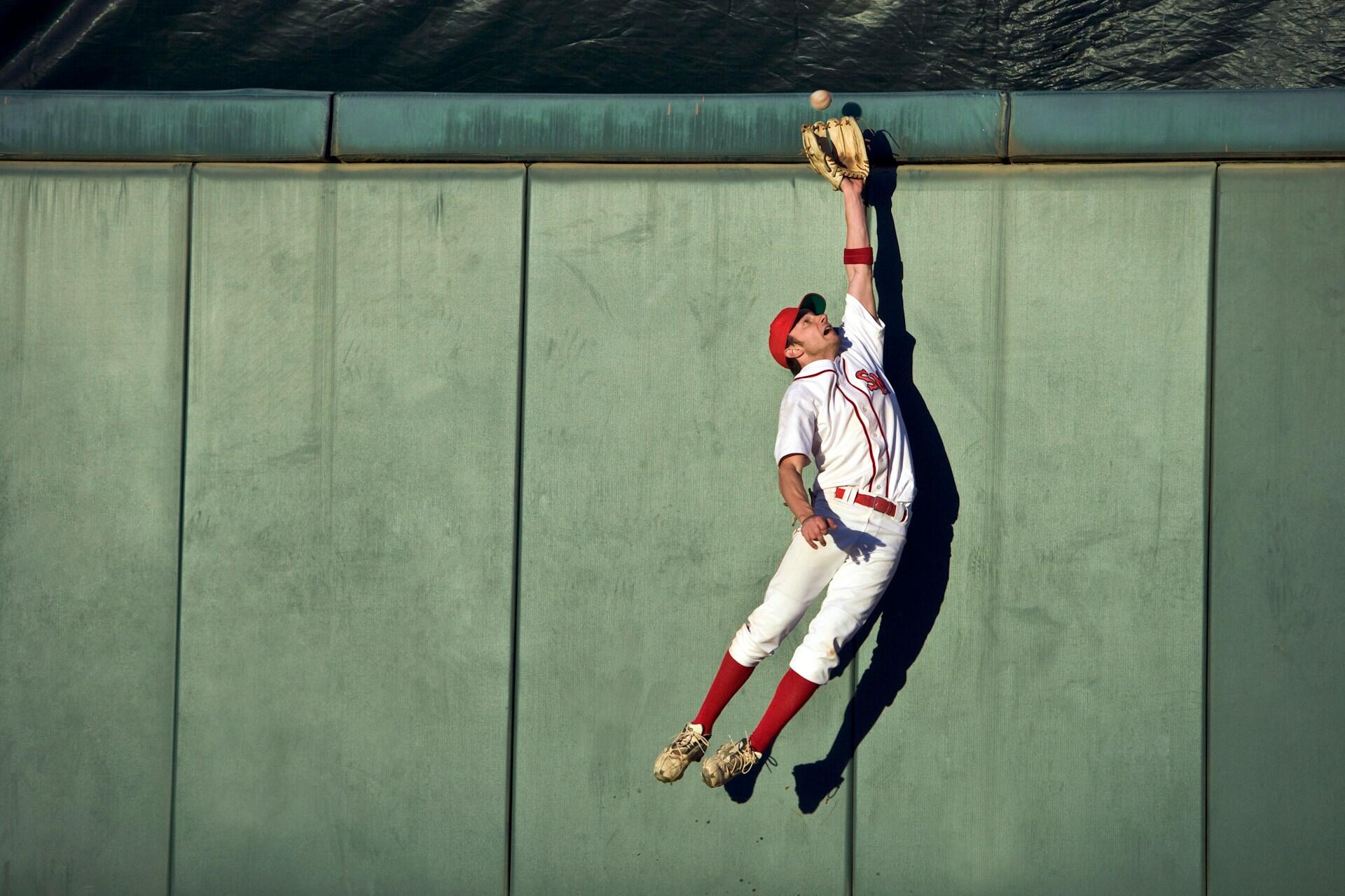 Un jugador de béisbol con uniforme blanco salta contra una pared verde, tratando de atrapar una bola alta con tu guante extendido en alto.