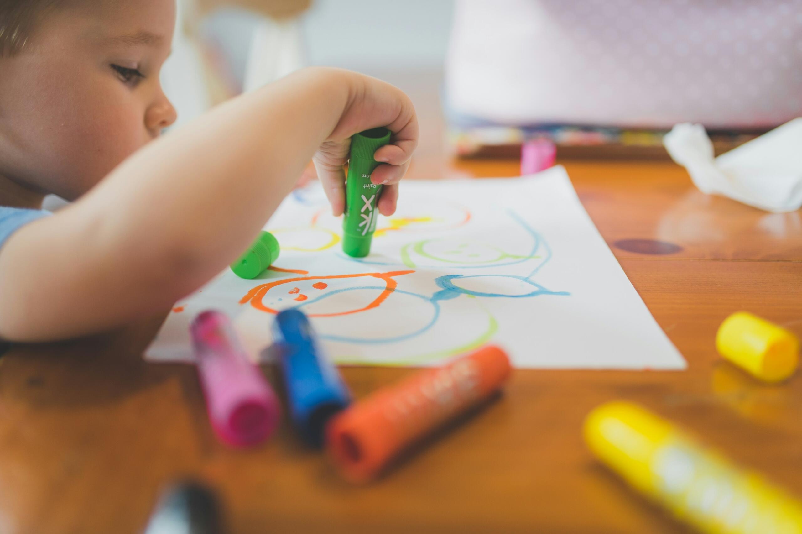 La mano de un niño sostiene un rotulador verde sobre un dibujo colorido en un papel, rodeado de otros rotuladores sobre una mesa de madera.
