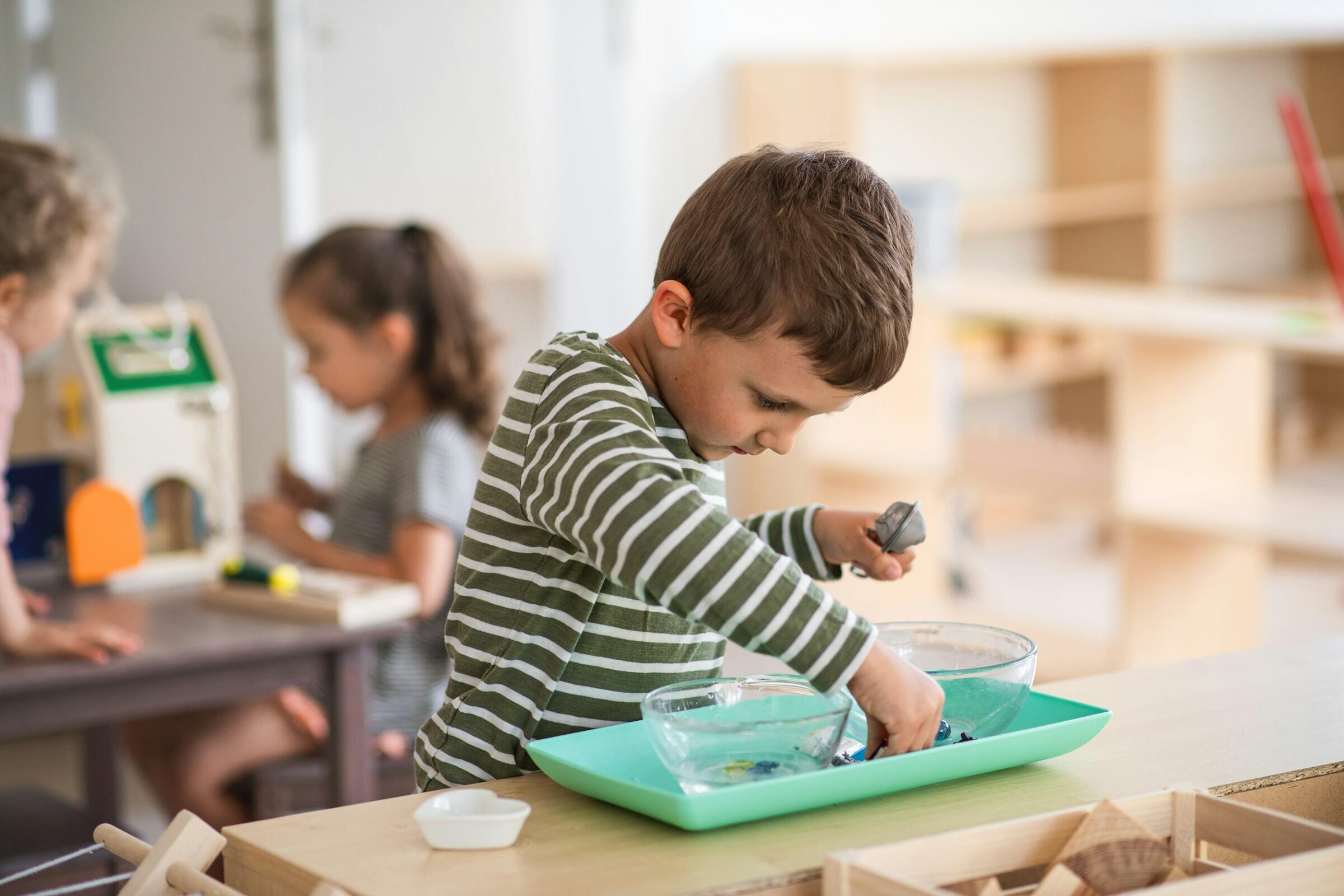 Un niño con una camiseta a rayas juega con objetos en un cuenco poco profundo lleno de agua sobre una mesa de madera en un aula luminosa.