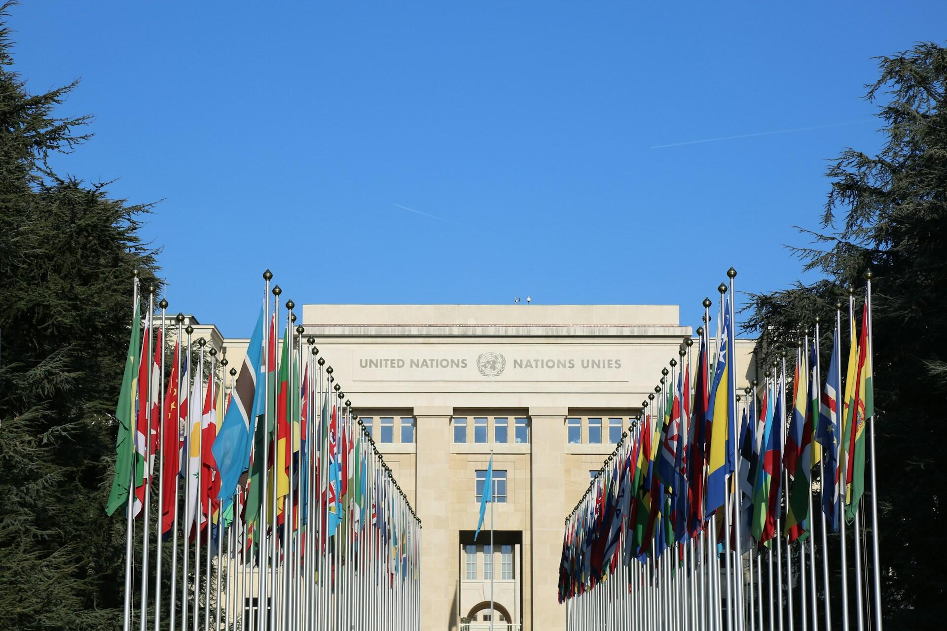 El edificio de las Naciones Unidas en Ginebra, rodeado por numerosas banderas de diferentes países contra un cielo azul claro.
