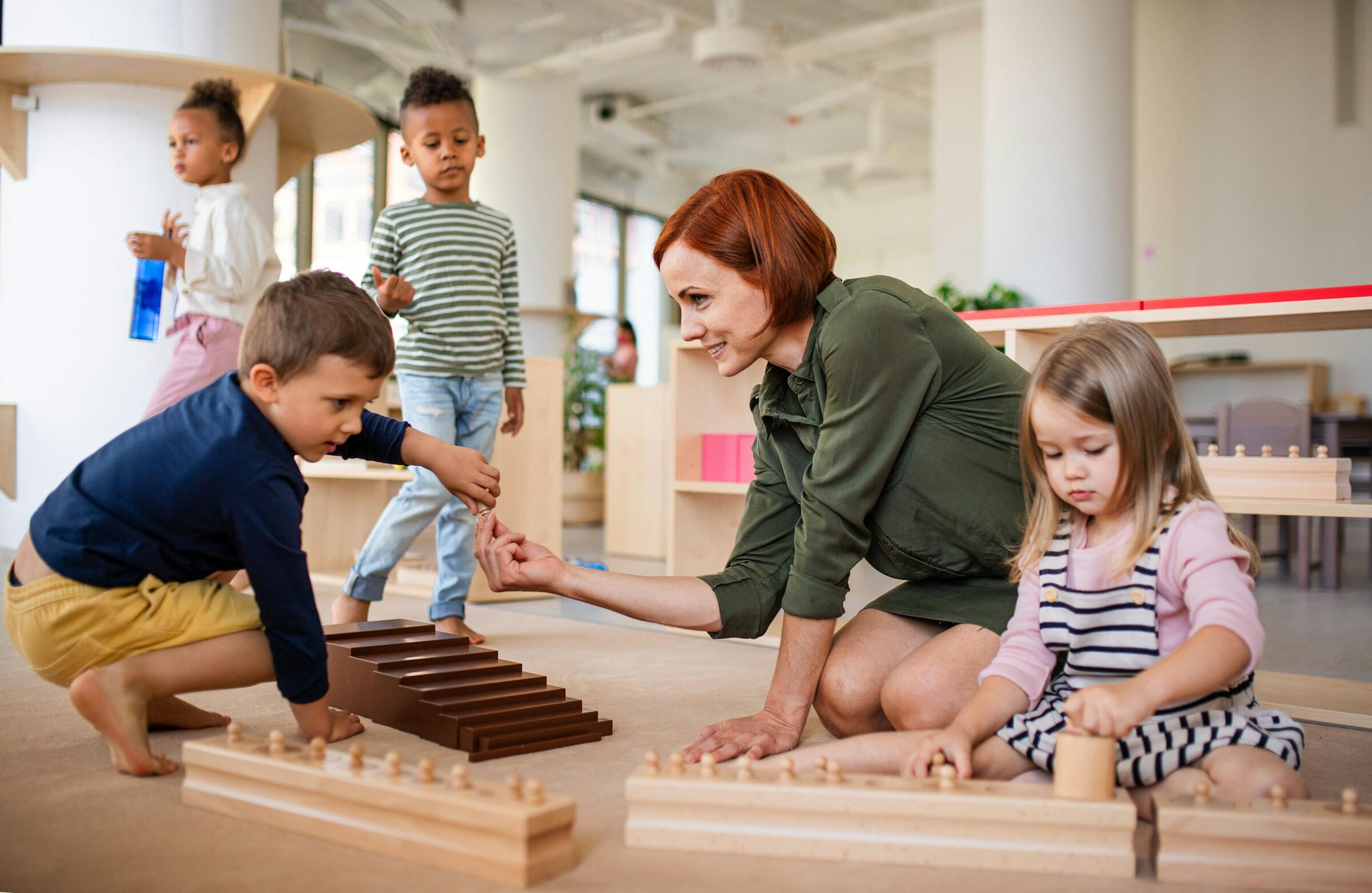 Un profesor interactúa con niños pequeños en un aula, jugando con juguetes de madera y materiales educativos en el suelo, la escalera de madera y los cilindros de montessori.