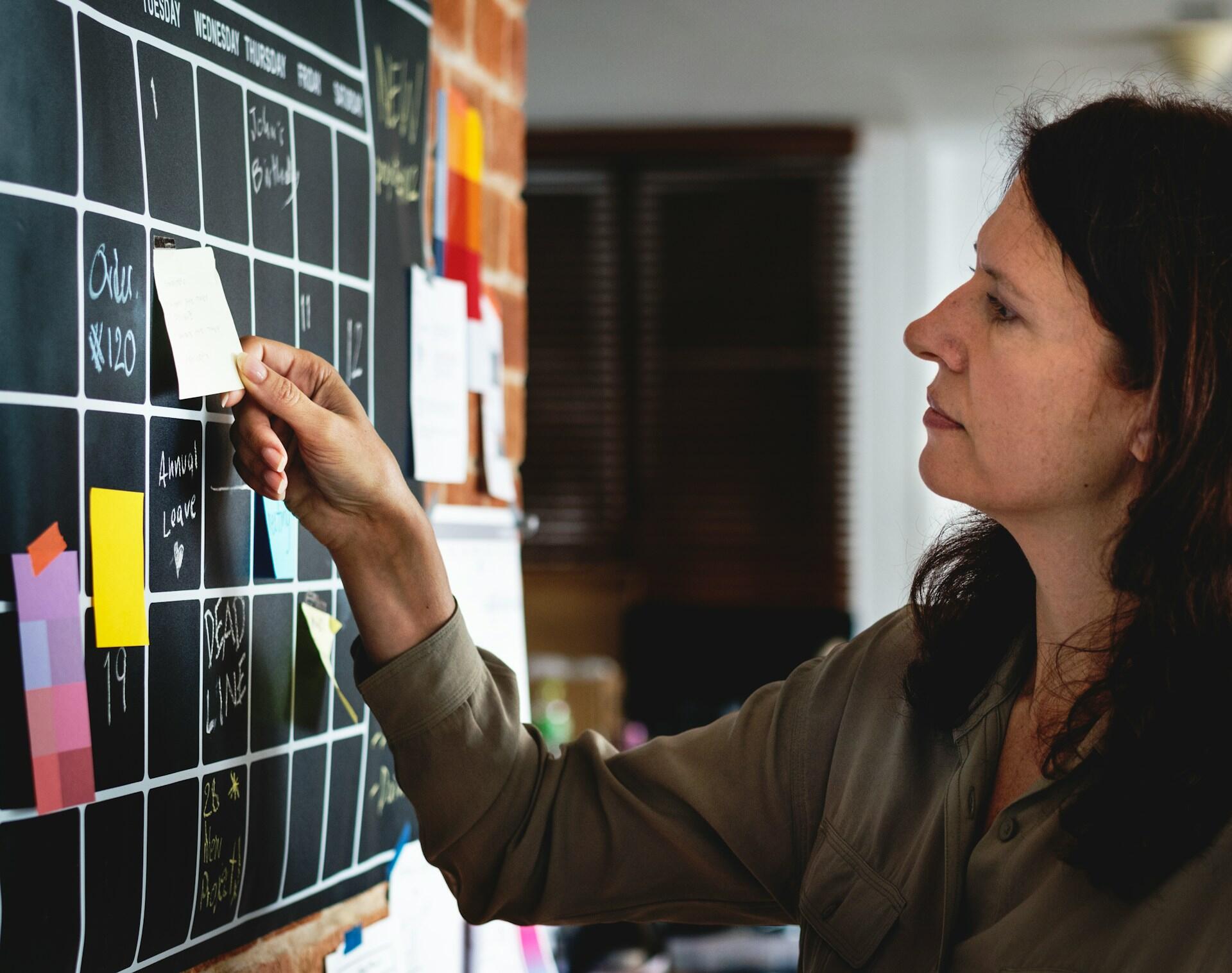 Mujer organizando tareas en un calendario mural. Usa notas adhesivas sobre una pizarra con cuadrícula semanal y textos escritos con tiza, como "DEADLINE" y "Annual Leave". Ambiente de oficina, planificación y gestión de proyectos.
