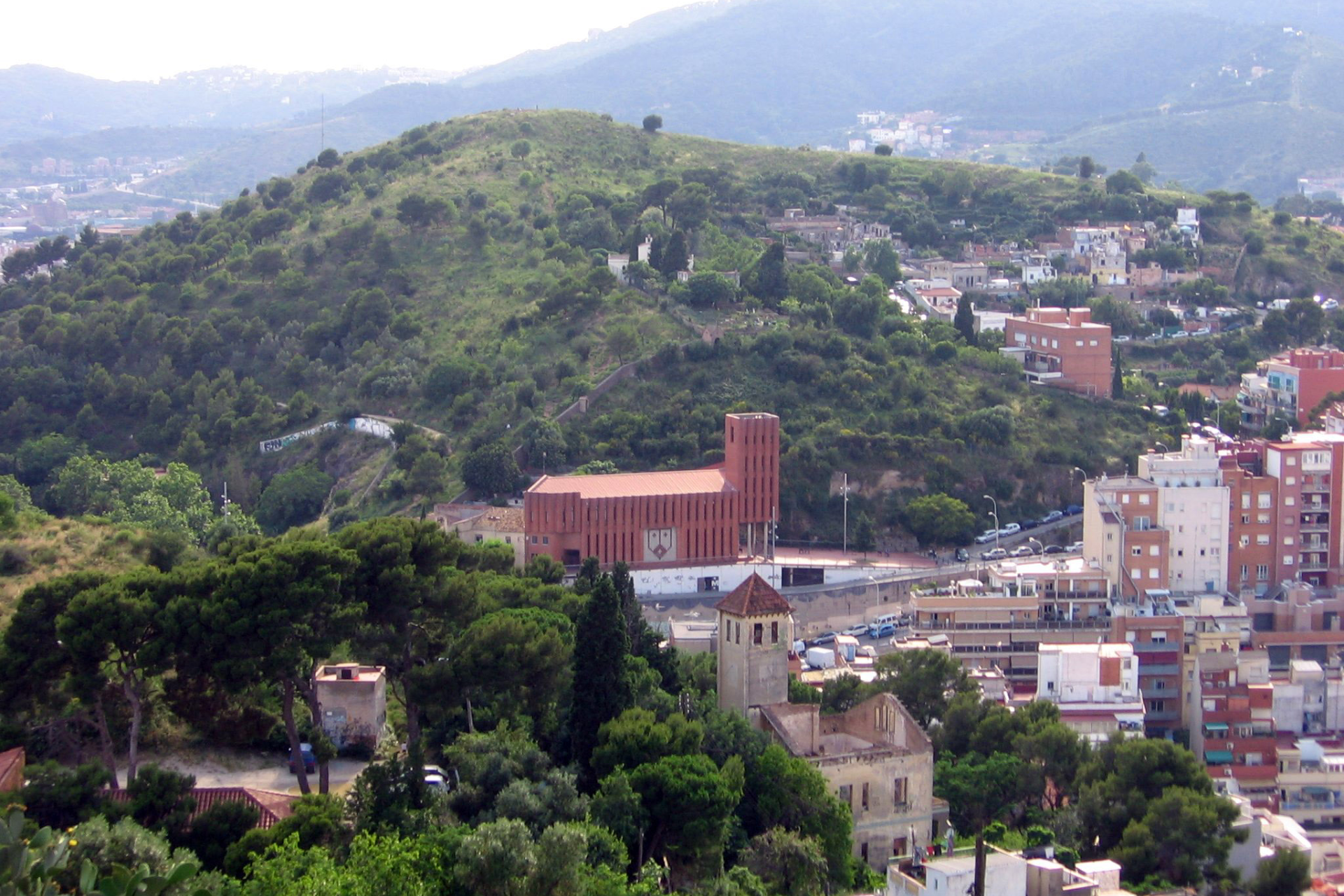 Vista de un paisaje montañoso con vegetación verde y una moderna iglesia de ladrillo rojo rodeada de edificios residenciales.