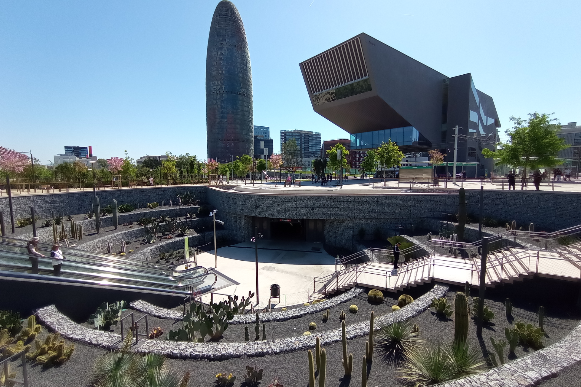 Un moderno parque urbano cuenta con un jardín de cactus, una escalera mecánica de cristal y una arquitectura contemporánea, con un cielo azul claro sobre tu.