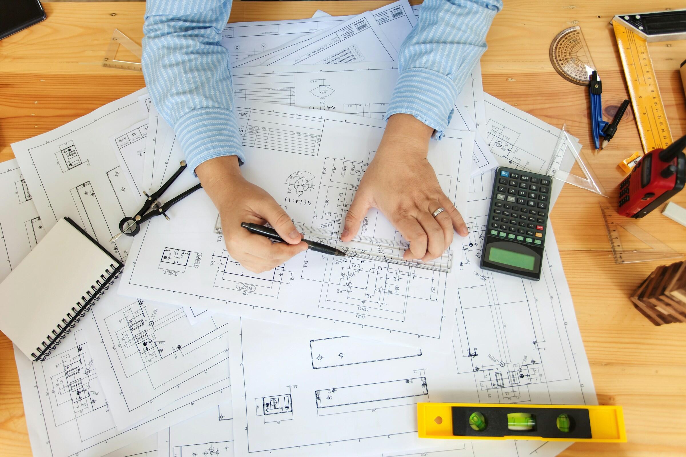 Las manos de una persona dibujando bocetos sobre planos rodeadas de una calculadora, herramientas de dibujo y planos de diseño sobre una mesa de madera.