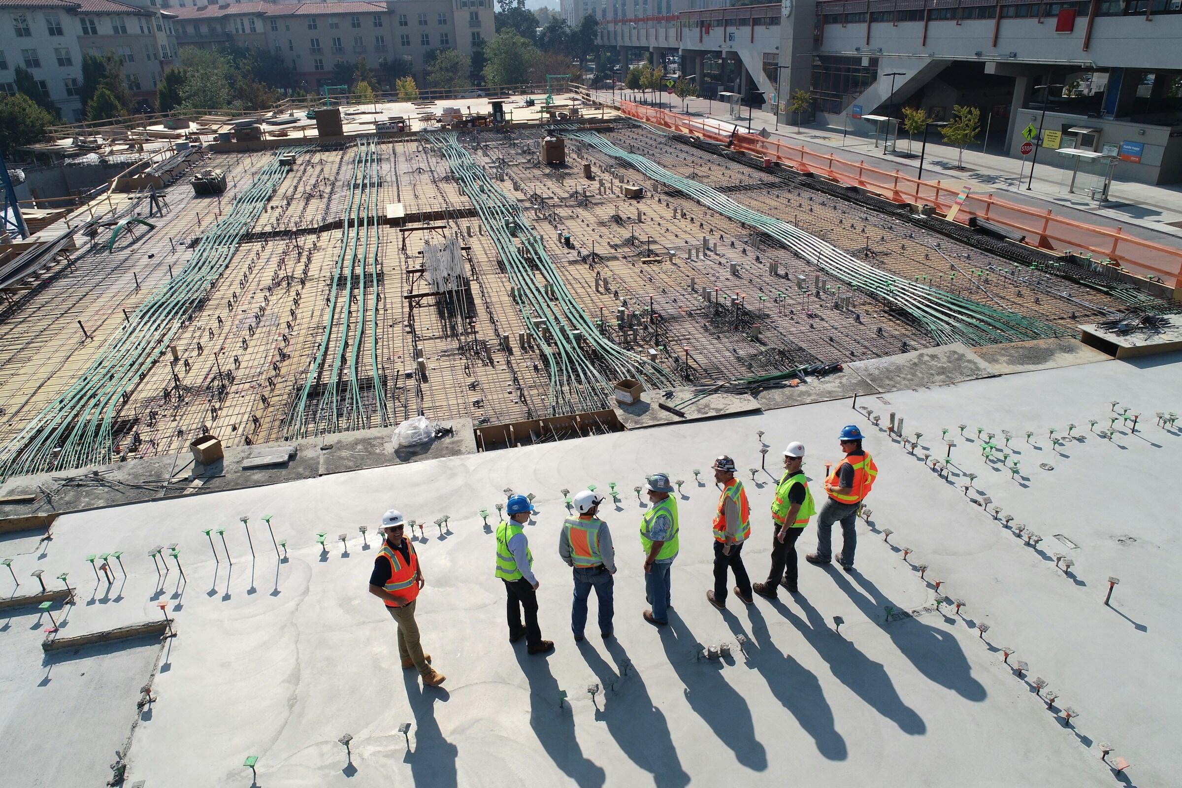 Un grupo de obreros de la construcción con chalecos de seguridad discute los planes sobre una gran losa de hormigón, supervisando una extensa obra.
