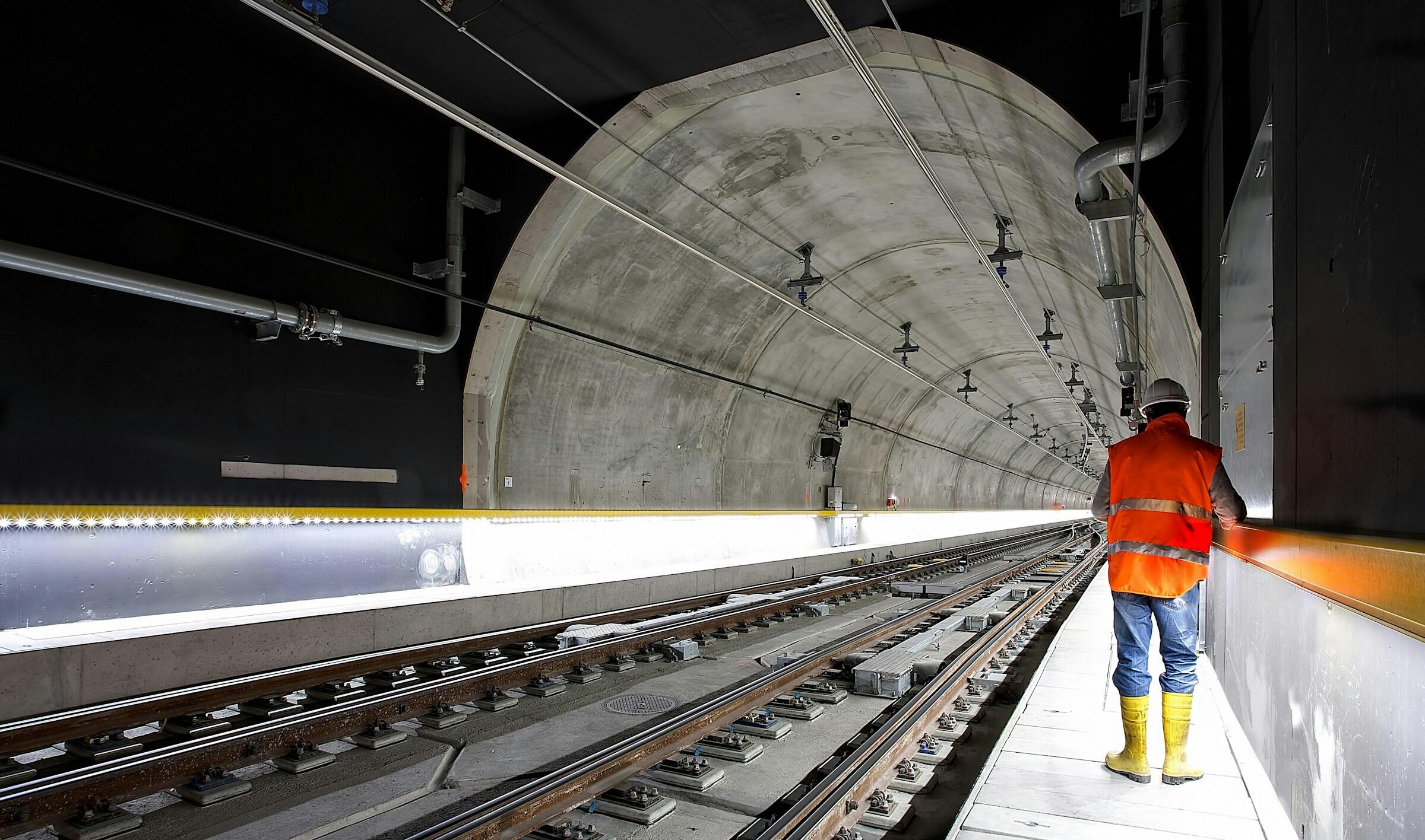 Un trabajador de la construcción con un chaleco naranja y un casco se encuentra junto a las vías del tren dentro de un túnel poco iluminado con un arco de hormigón.