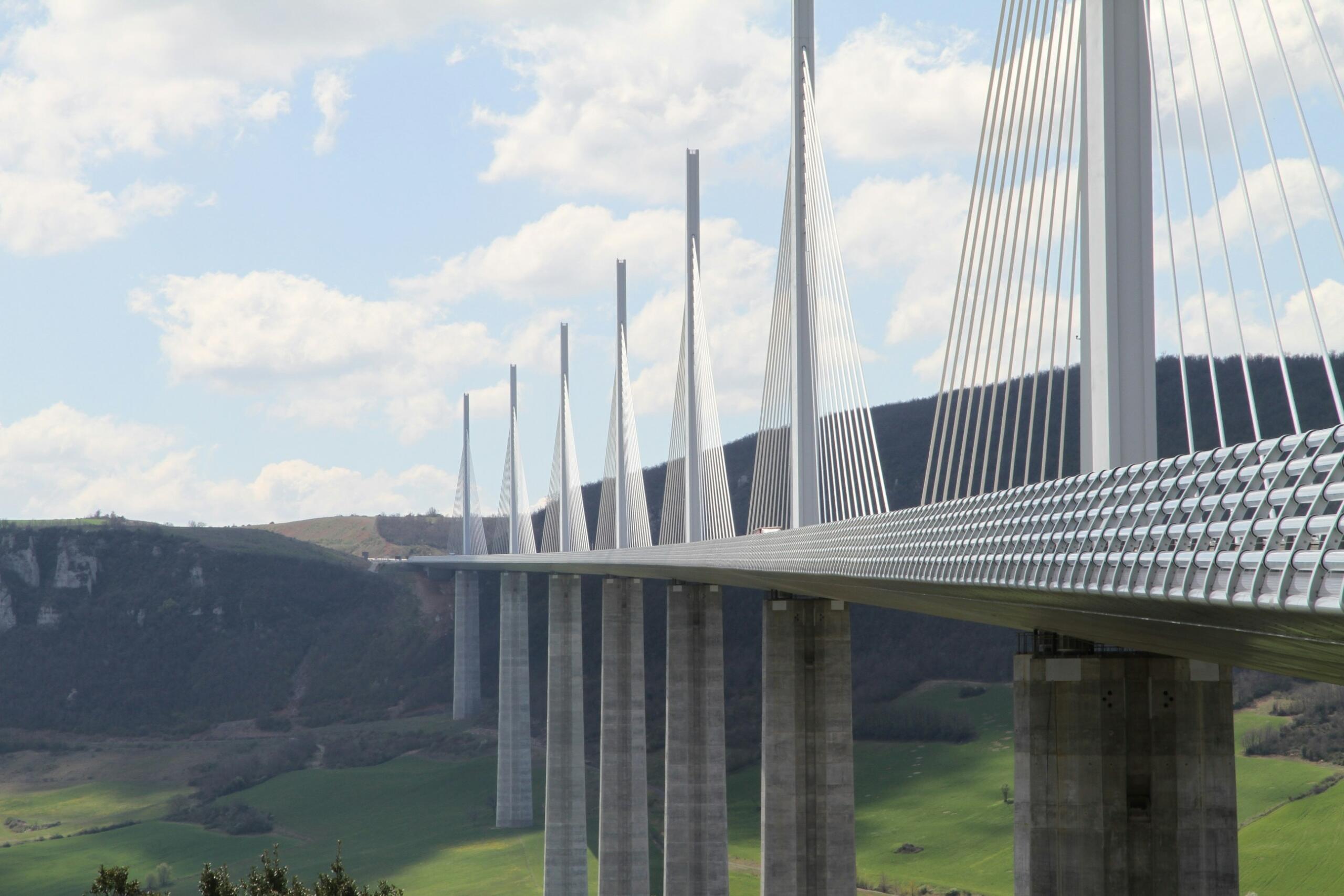 Una impresionante vista del viaducto de Millau, con sus altos y elegantes pilones y tirantes, rodeado de exuberantes colinas verdes bajo un cielo azul.