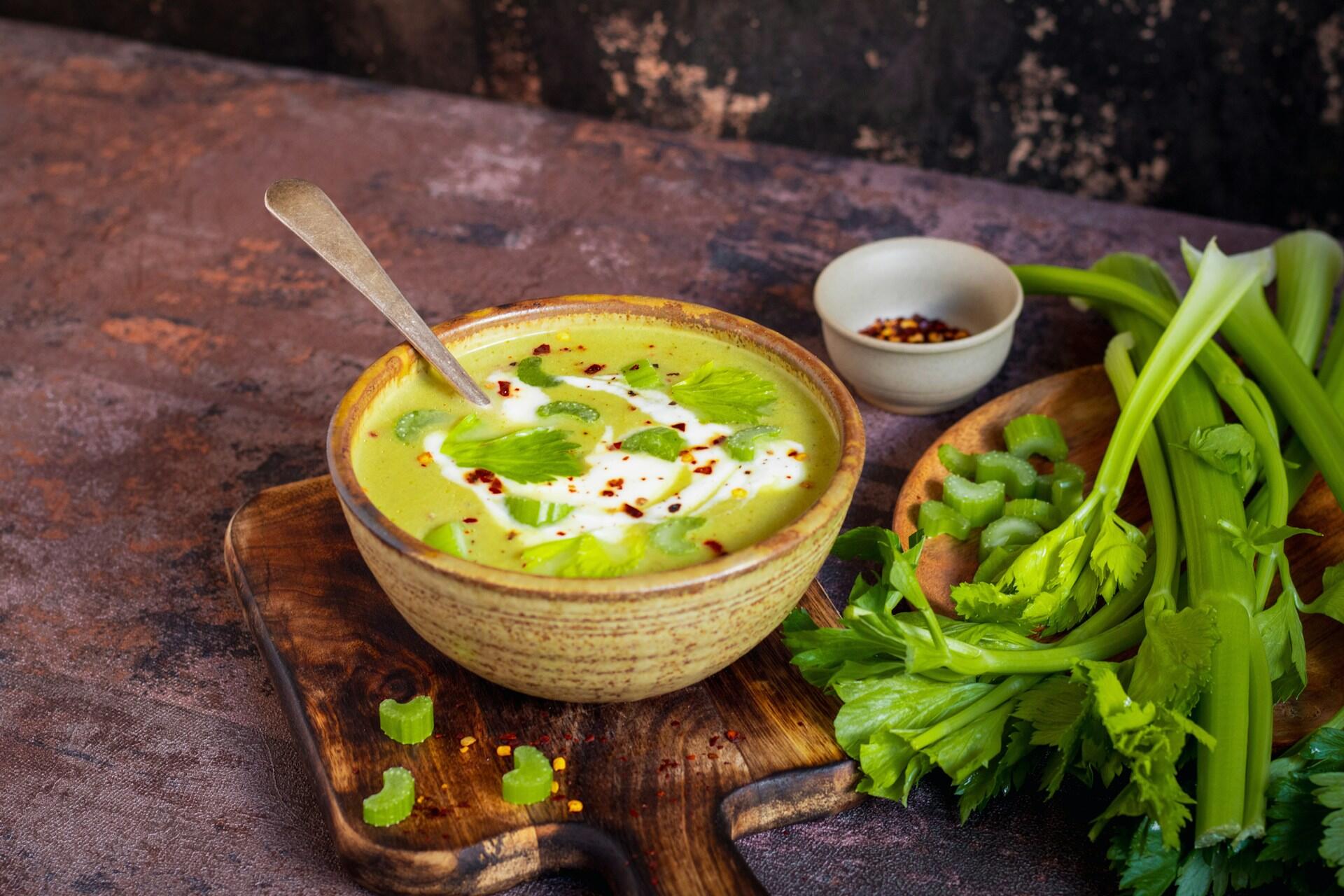 Sopa cremosa de apio servida en tazón rústico sobre tabla de madera. Decorada con crema, hojas de apio y chile en hojuelas. Acompañada de tallos de apio frescos y especias en fondo oscuro. Estilo casero y saludable.