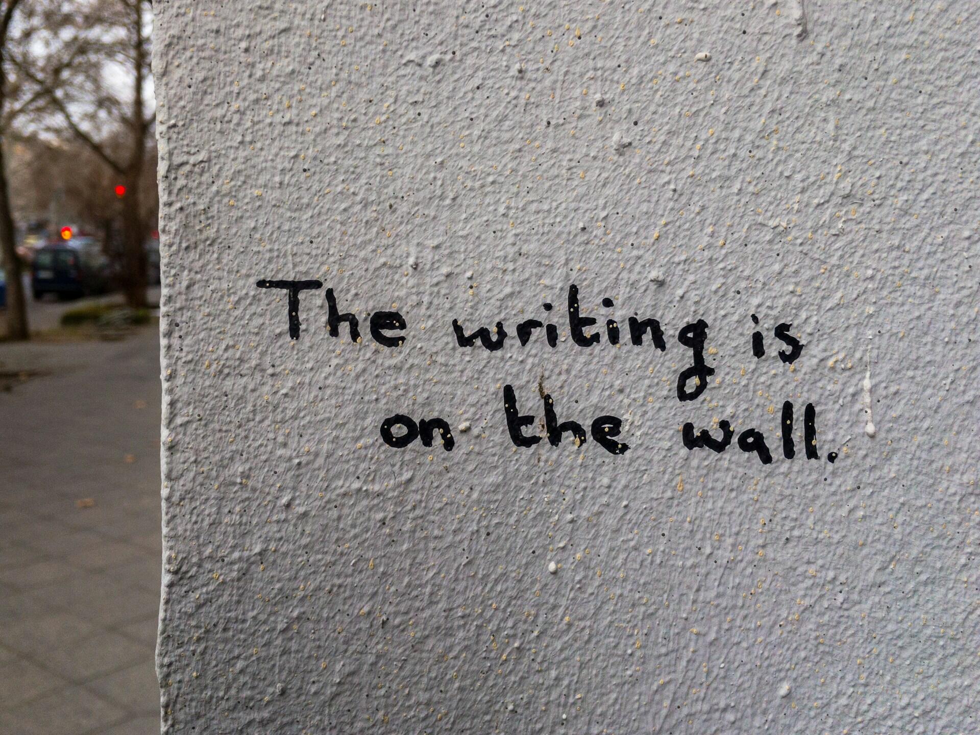 En una pared blanca con textura hay un grafiti que dice: «La escritura está en la pared» en inglés, con una escena callejera borrosa al fondo.