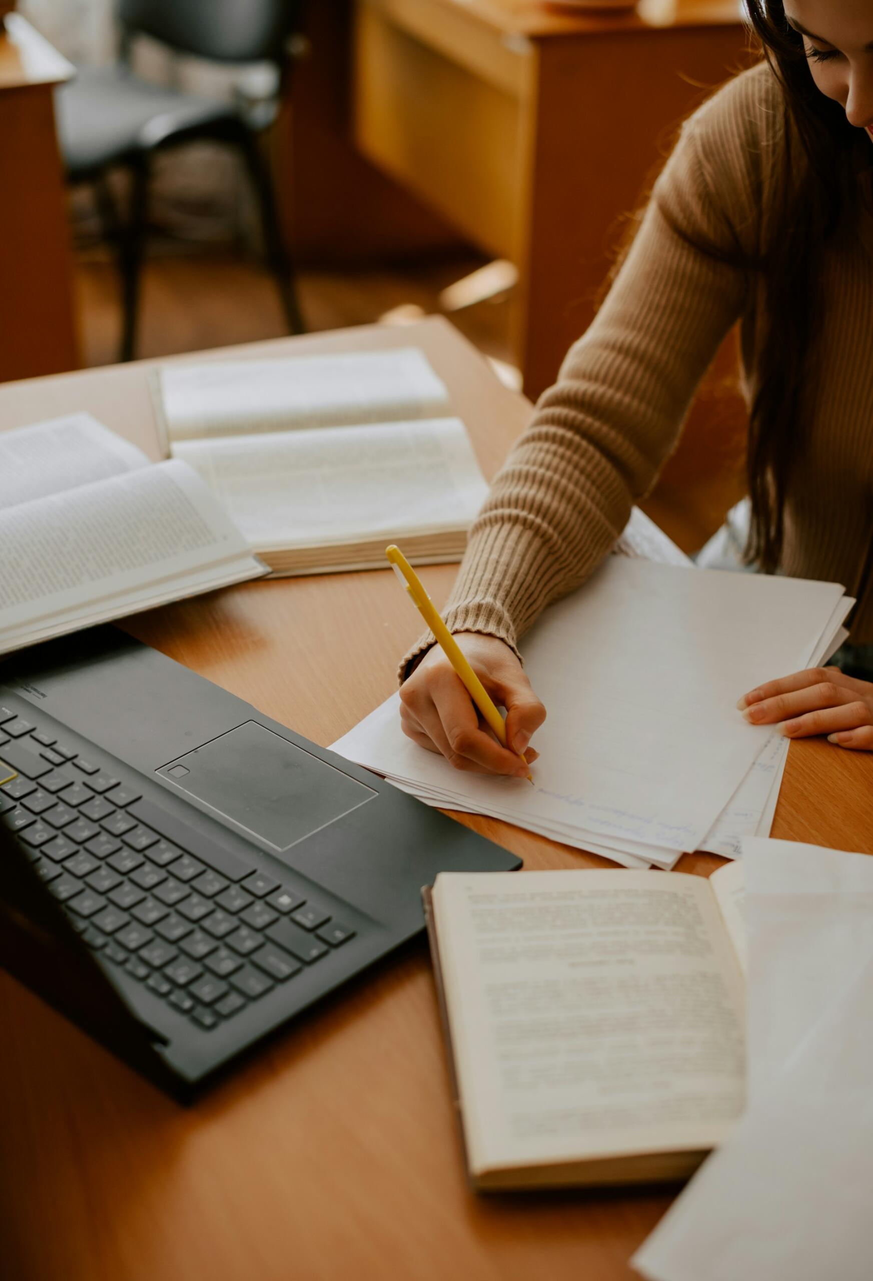 Un estudiante escribe notas con un lápiz amarillo en un papel en blanco, rodeado de libros abiertos y un ordenador portátil sobre una mesa de madera.