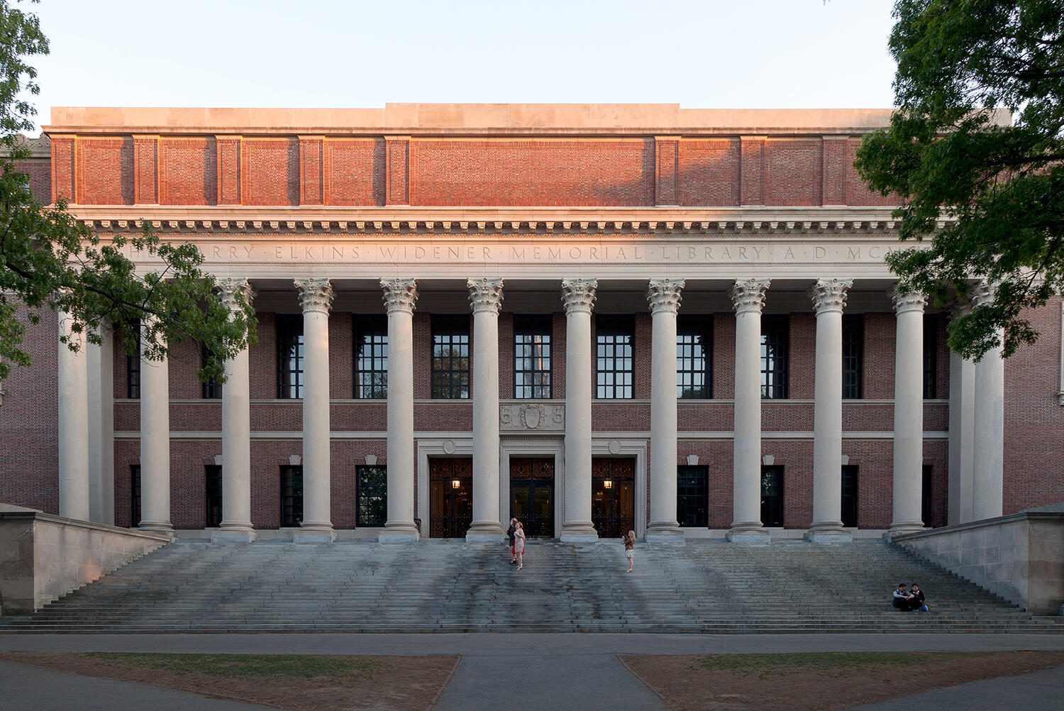 Vista frontal de la Biblioteca Harry Elkins Widener Memorial, en la que se aprecia su gran escalera y sus columnas clásicas al atardecer. Hay estudiantes presentes.
