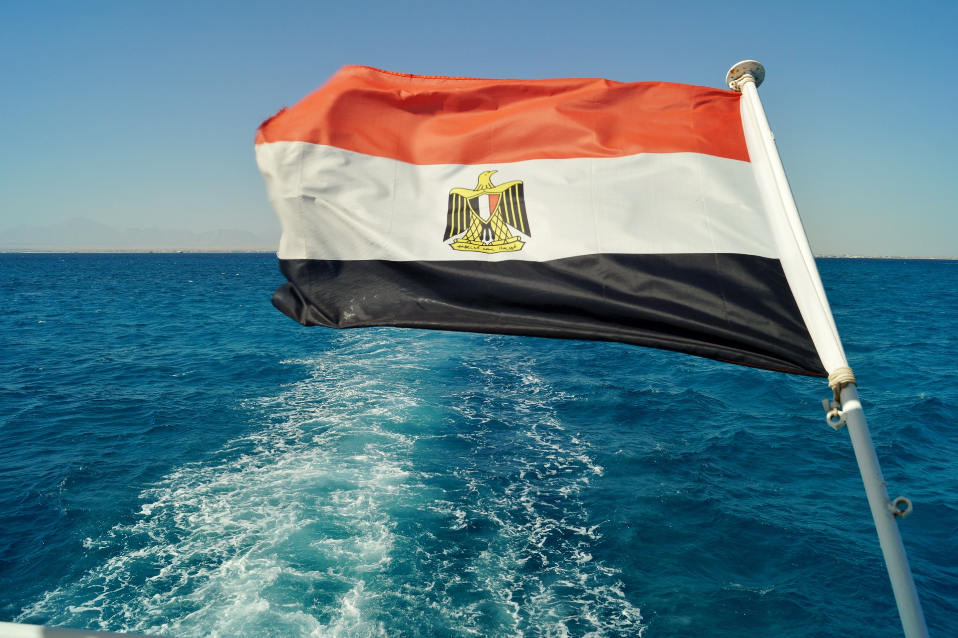 Bandera de Egipto con tres franjas horizontales de colores rojo, blanco y negro, y el águila dorada de Saladin en el centro.