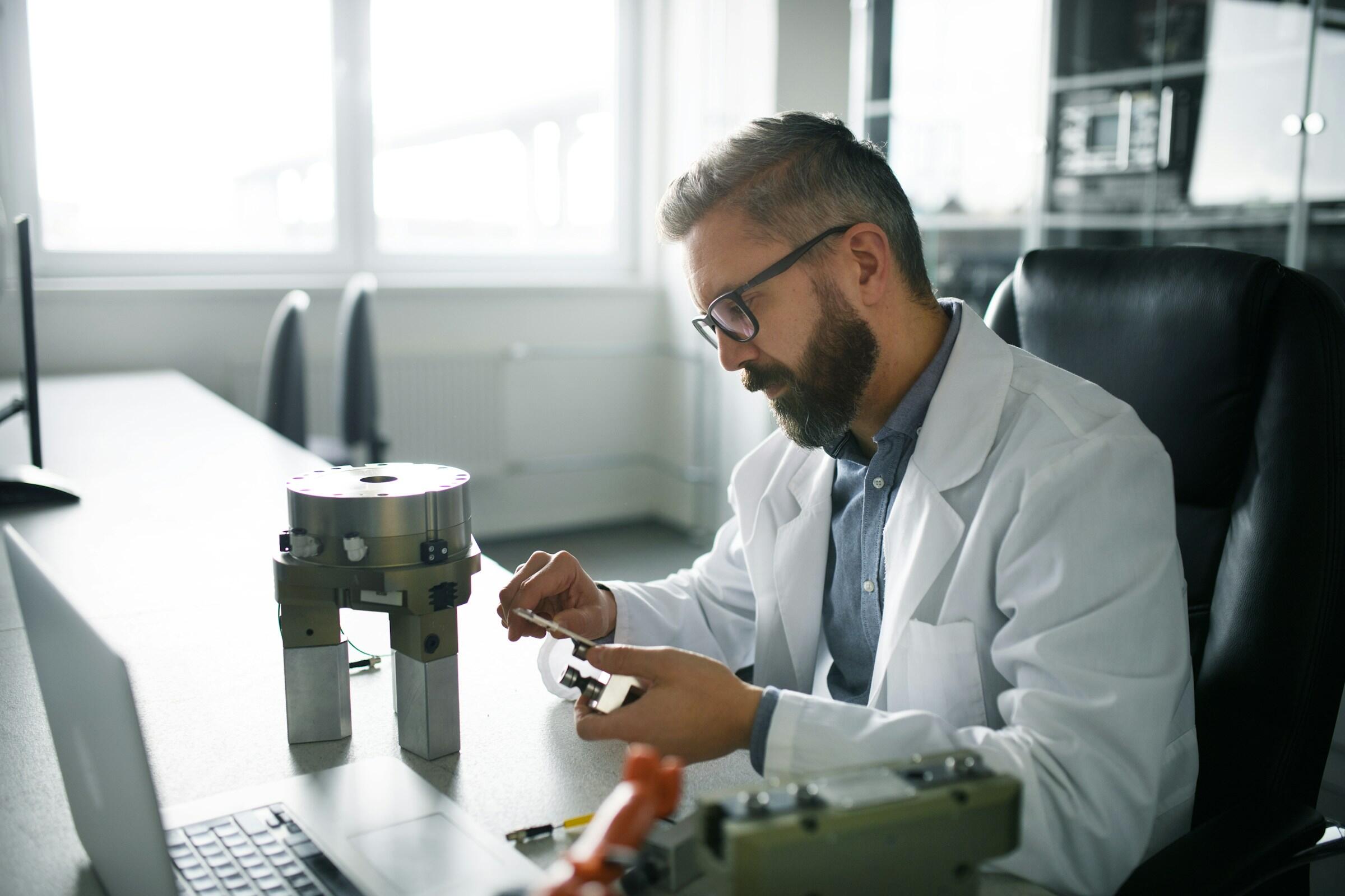 Una persona con bata blanca de laboratorio examina un componente mecánico en un escritorio, con un ordenador portátil y otras herramientas cerca. La luz natural entra por las ventanas.