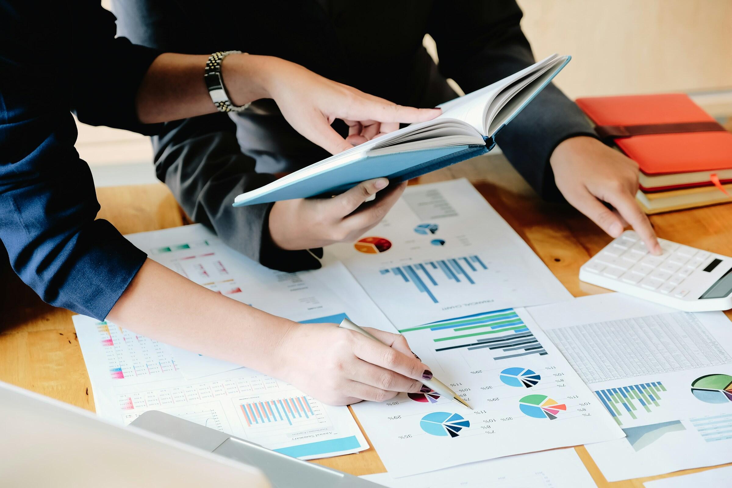 Dos profesionales analizan documentos con tablas y gráficos, discutiendo los resultados mientras uno señala los datos y el otro sostiene un libro.