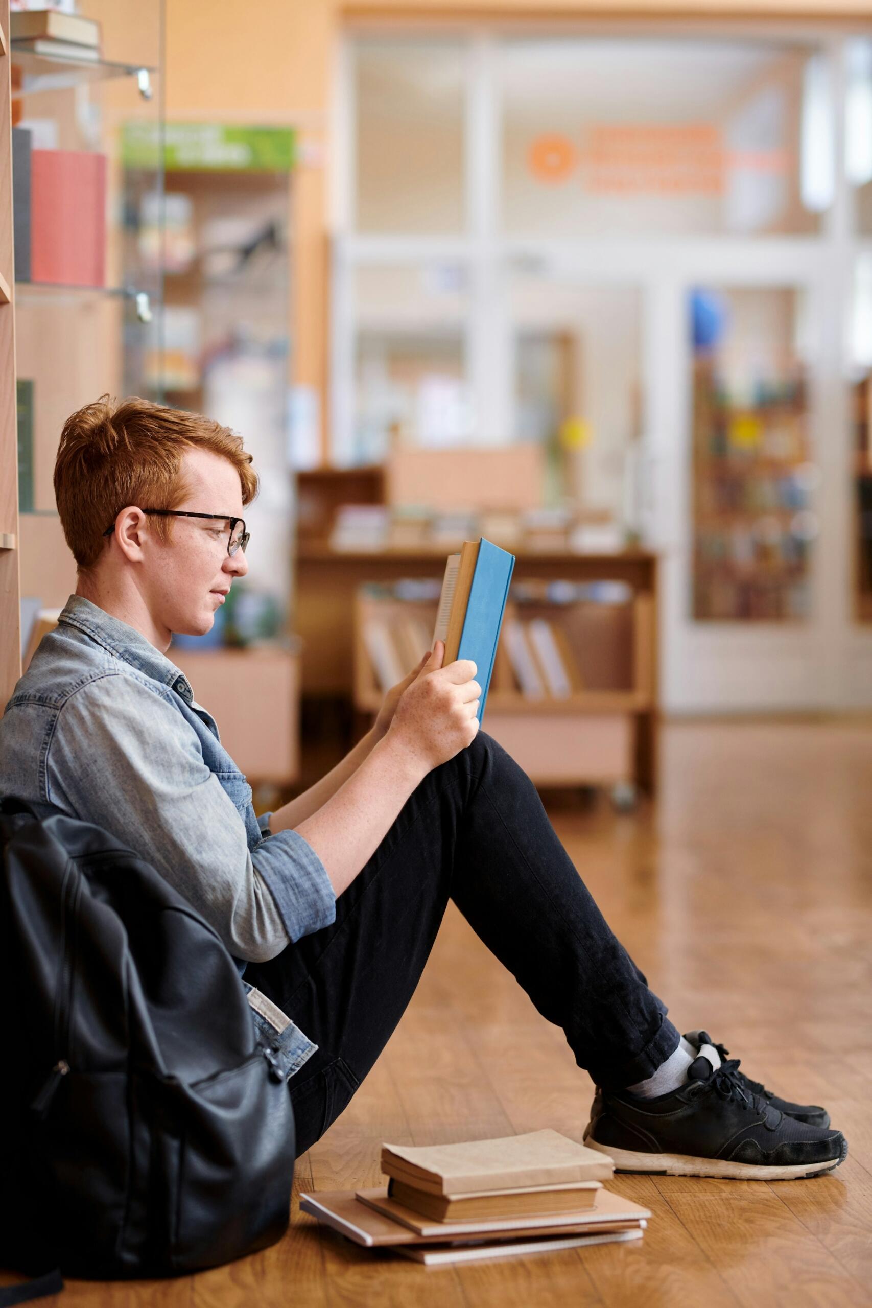 Un joven sentado en el suelo de una biblioteca, absorto en un libro, con una mochila y varios libros apilados a su lado.