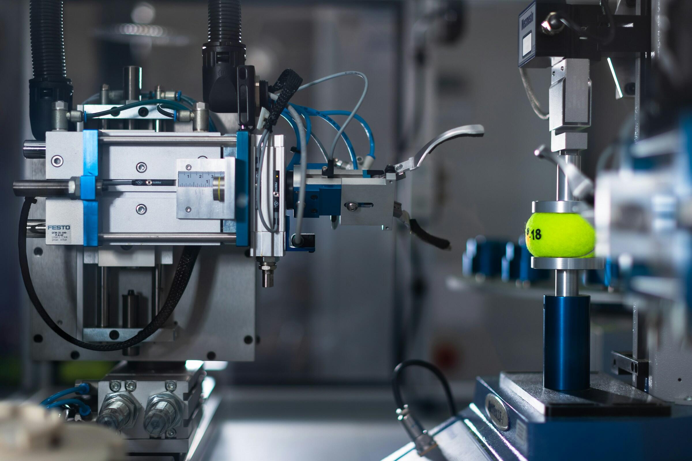 Primer plano de una máquina automatizada sujetando una pelota de tenis amarilla, en el que se aprecian los componentes y controles de ingeniería de precisión.