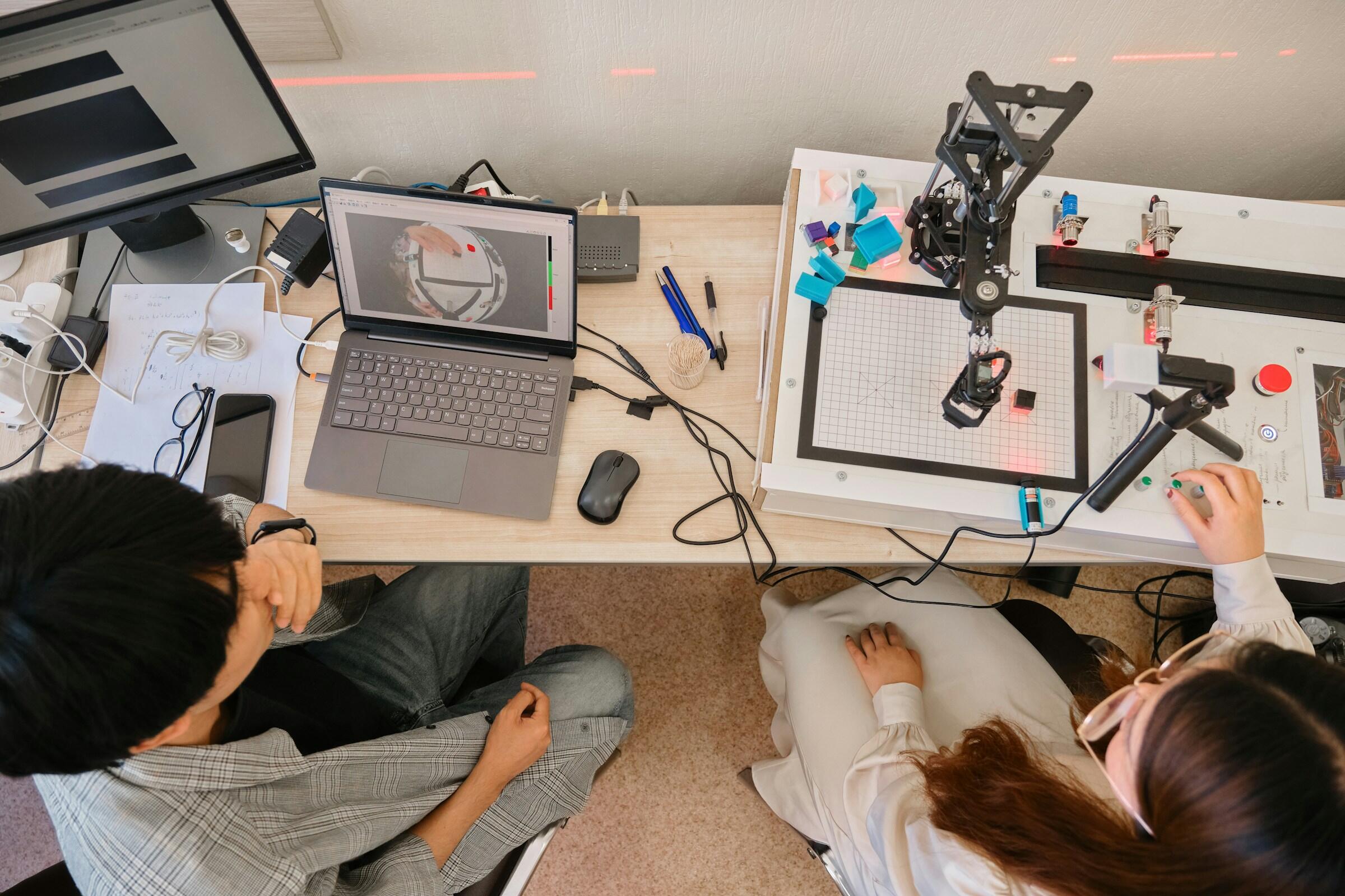Dos investigadores trabajando en un proyecto de robótica, utilizando ordenadores portátiles y un brazo robótico, rodeados de herramientas y papeles sobre una mesa.
