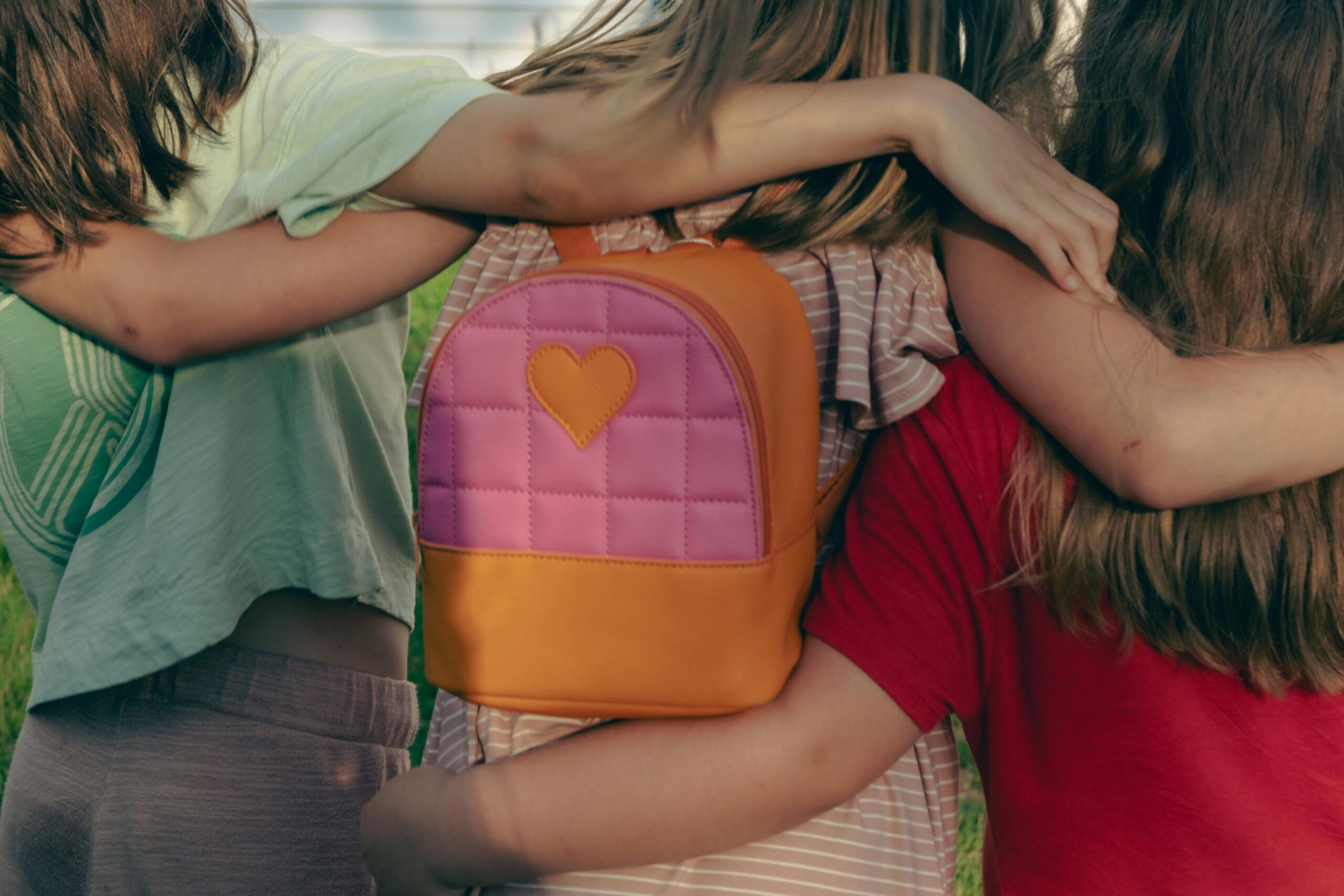 Tres niñas se abrazan entre sí, sosteniendo una mochila colorida. 