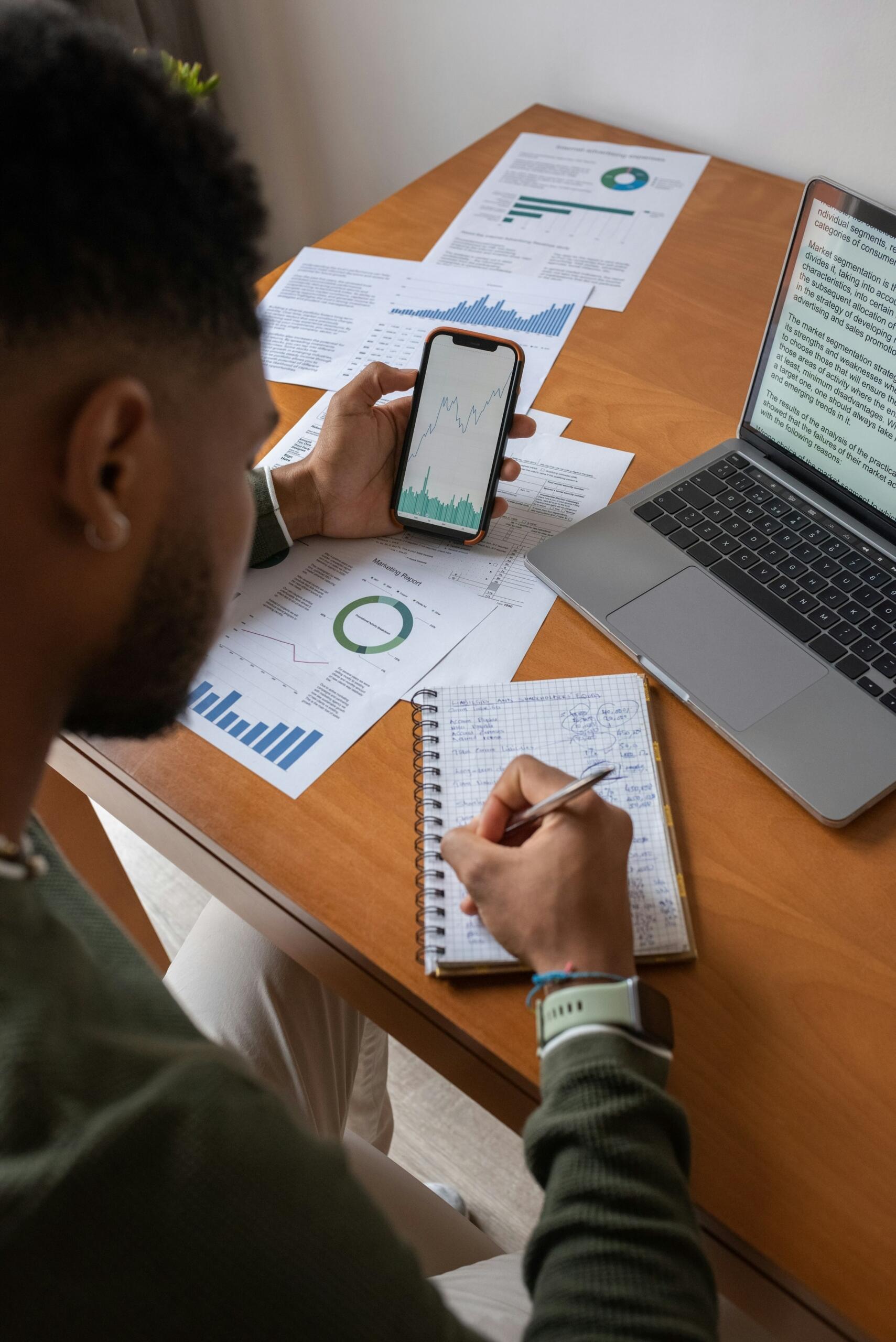 Una persona estudia datos financieros en un smartphone mientras toma notas en un cuaderno, rodeada de documentos sobre un escritorio de madera.