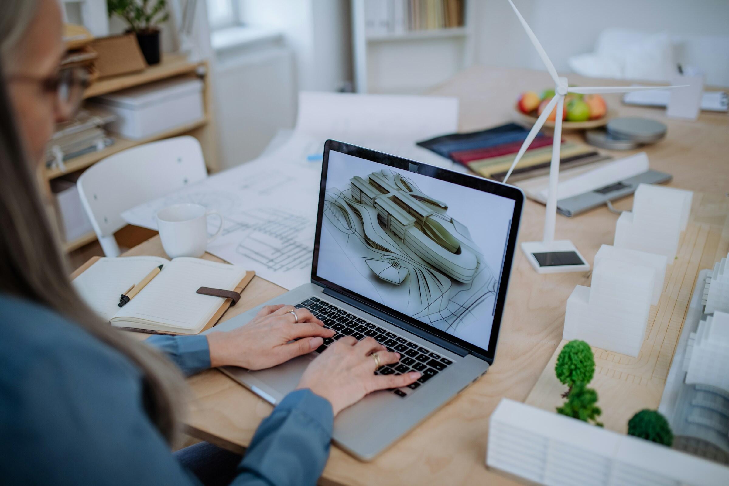 Una persona trabaja en un ordenador portátil en el que se muestra un modelo arquitectónico en 3D, rodeada de bocetos, un cuaderno y materiales de diseño sobre una mesa.