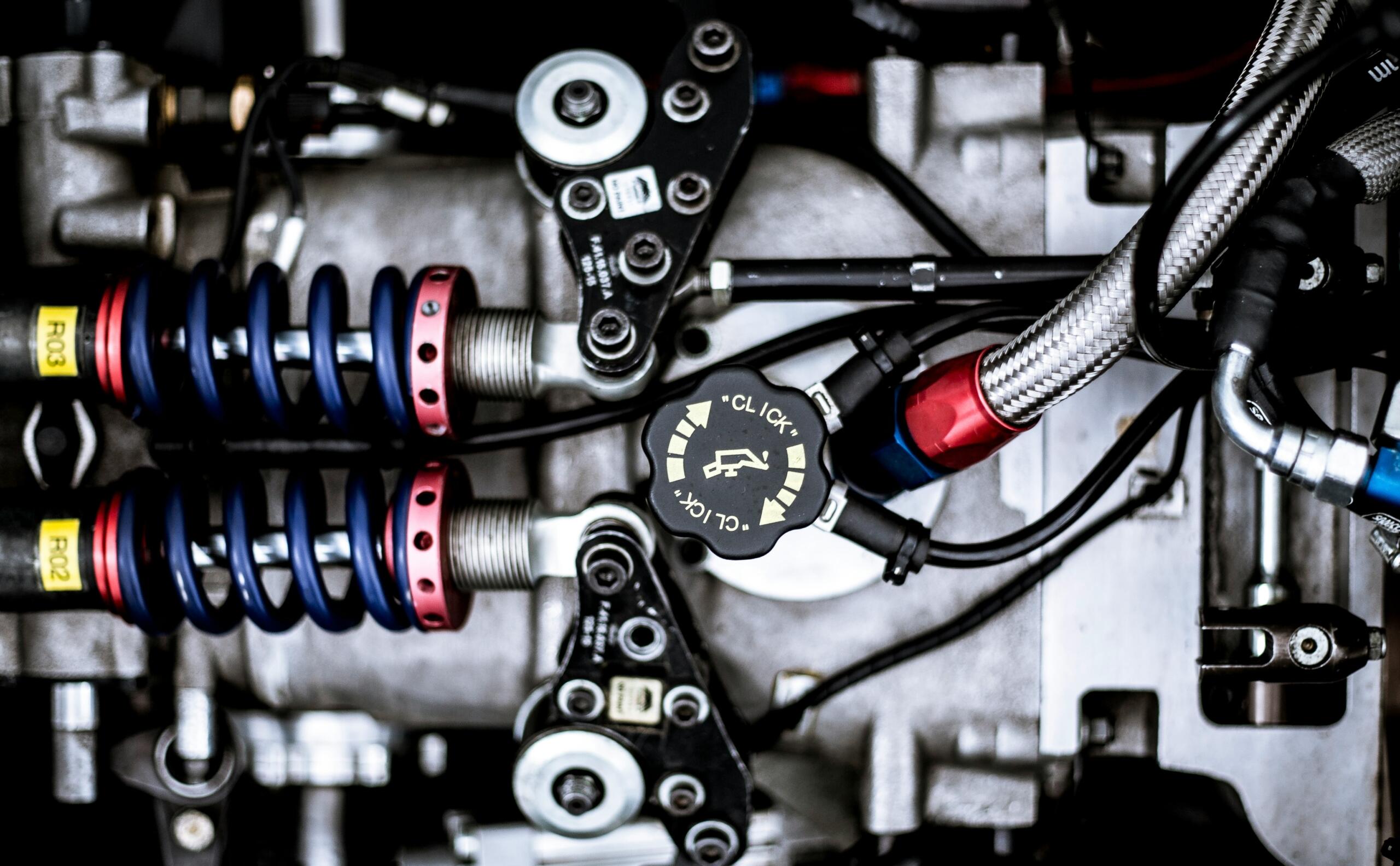 Close-up of a high-performance engine with blue and red suspension springs, complex tubing, and a detailed valve adjuster.