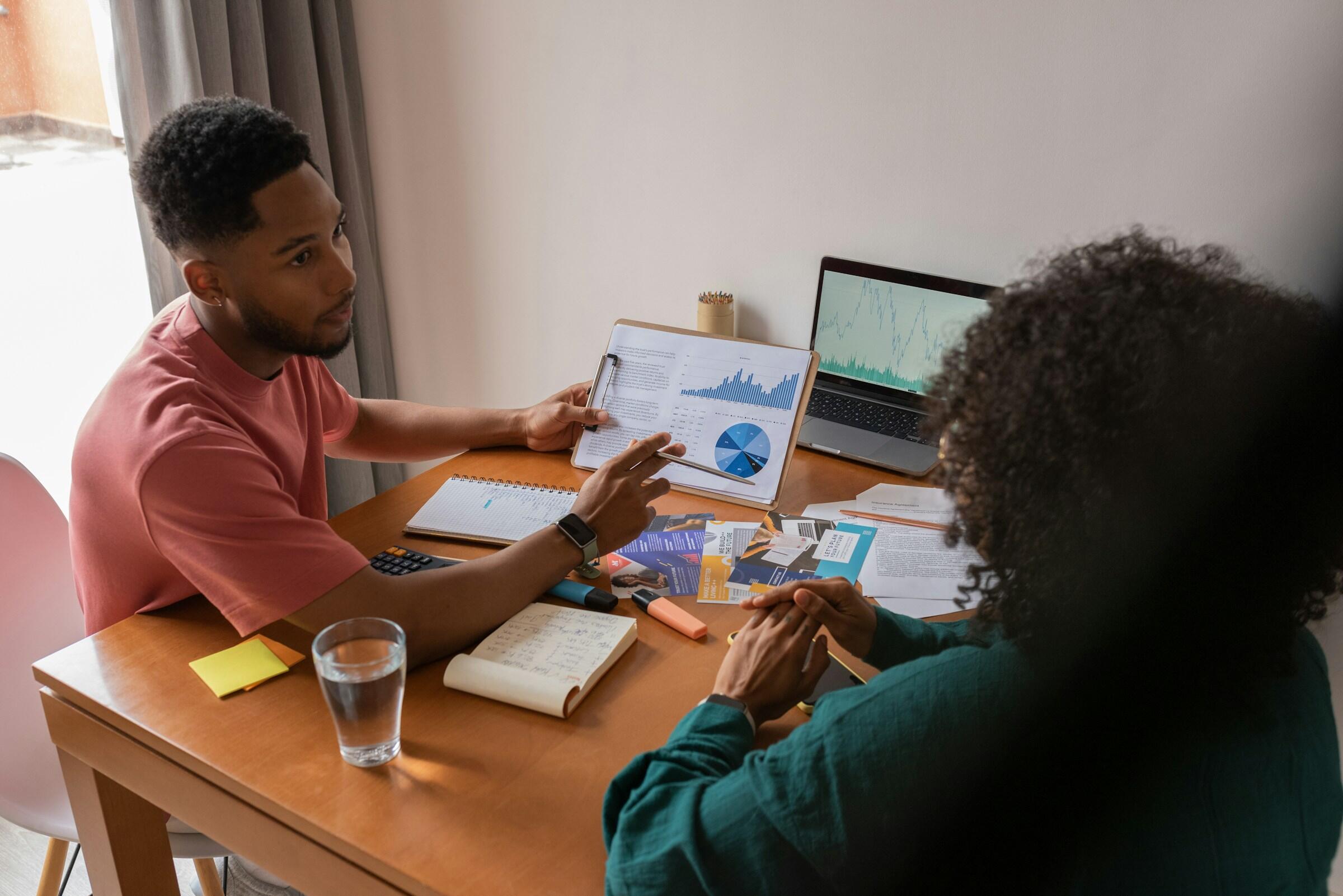  un hombre a la izquierda de la mesa que lleva una camiseta rosa. Tiene el cabello corto y barba, y está gesticulando hacia un documento o libreta que sostiene con la mano derecha. Su expresión sugiere que está explicando algo importante. Sobre la mesa frente a él hay una libreta abierta, una calculadora, un vaso de agua y algunas notas adhesivas. También se ve una computadora portátil en la mesa, mostrando lo que parece ser un gráfico o una tabla.