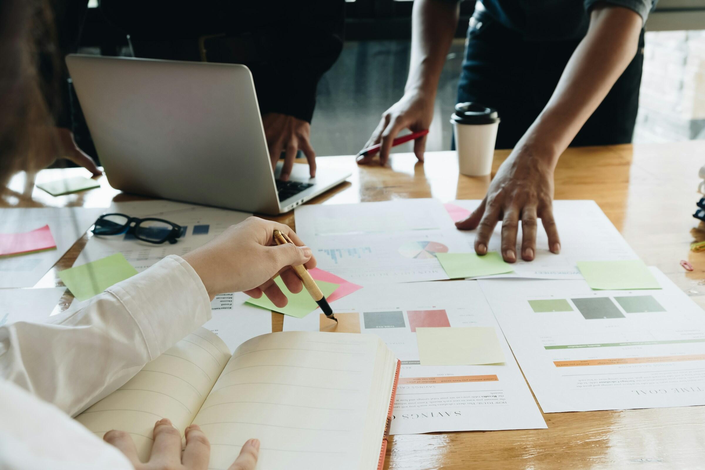 Un grupo de personas colabora en una mesa cubierta de papeles, notas adhesivas y un ordenador portátil, debatiendo activamente y tomando notas.