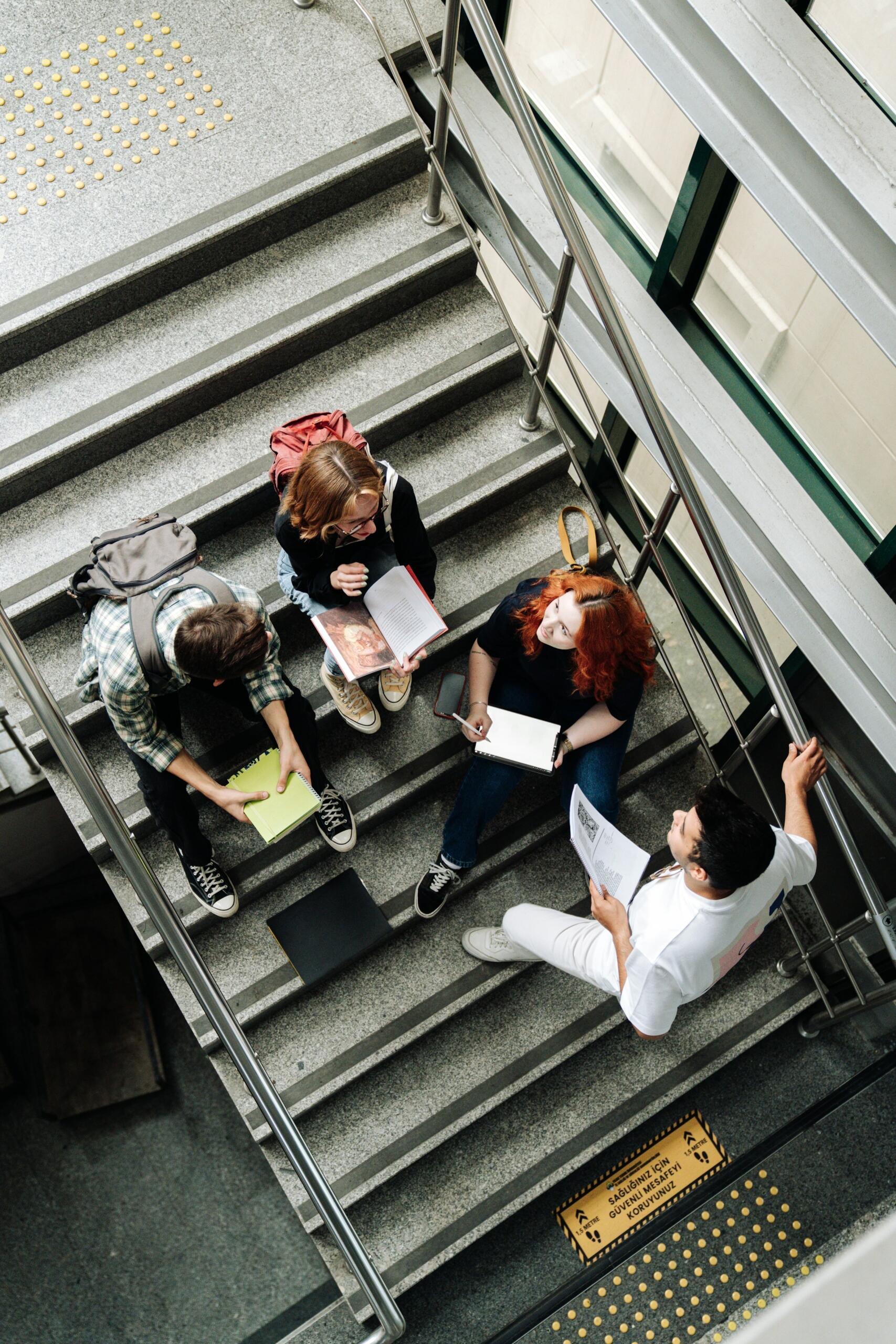 Un grupo de estudiantes está sentado en las escaleras estudiando juntos, rodeados de libros y apuntes en un edificio moderno.