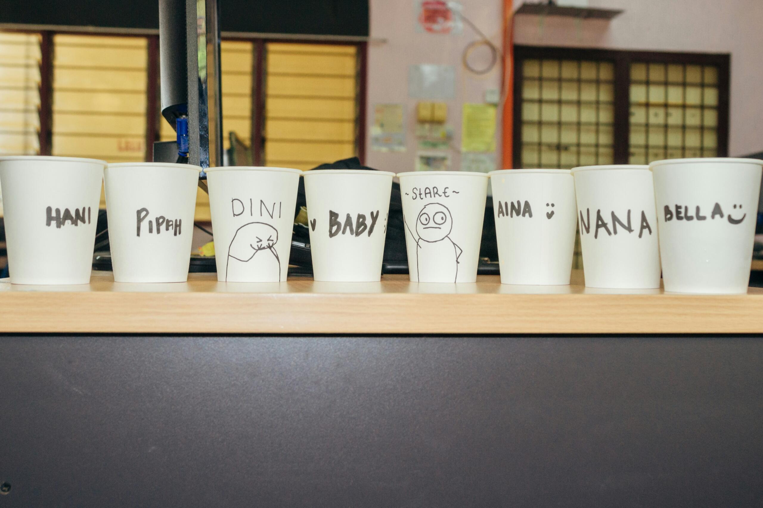 Una fila de vasos de papel blancos etiquetadas con nombres y dibujos sencillos, entre ellos «HANI», «PIPAH», «DINI», «BABY» y otros, colocadas sobre una mesa.