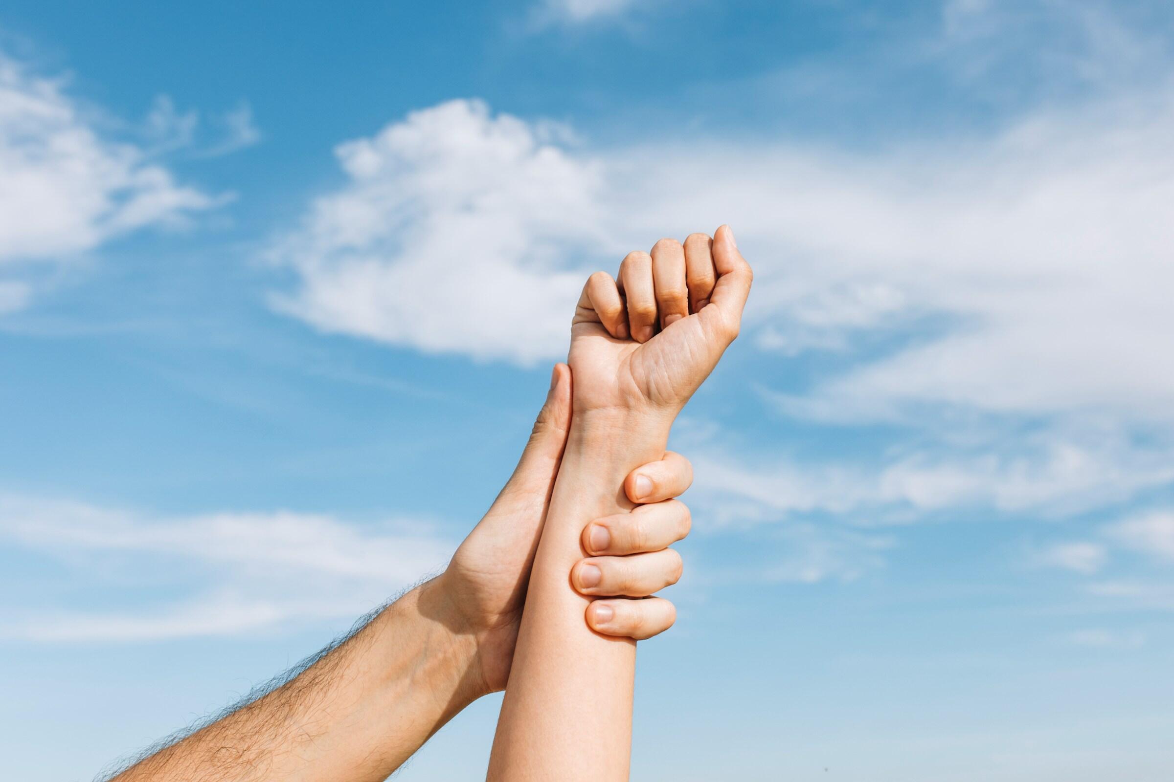 Dos manos entrelazadas contra un cielo azul brillante con nubes esponjosas, simbolizando conexión o apoyo.