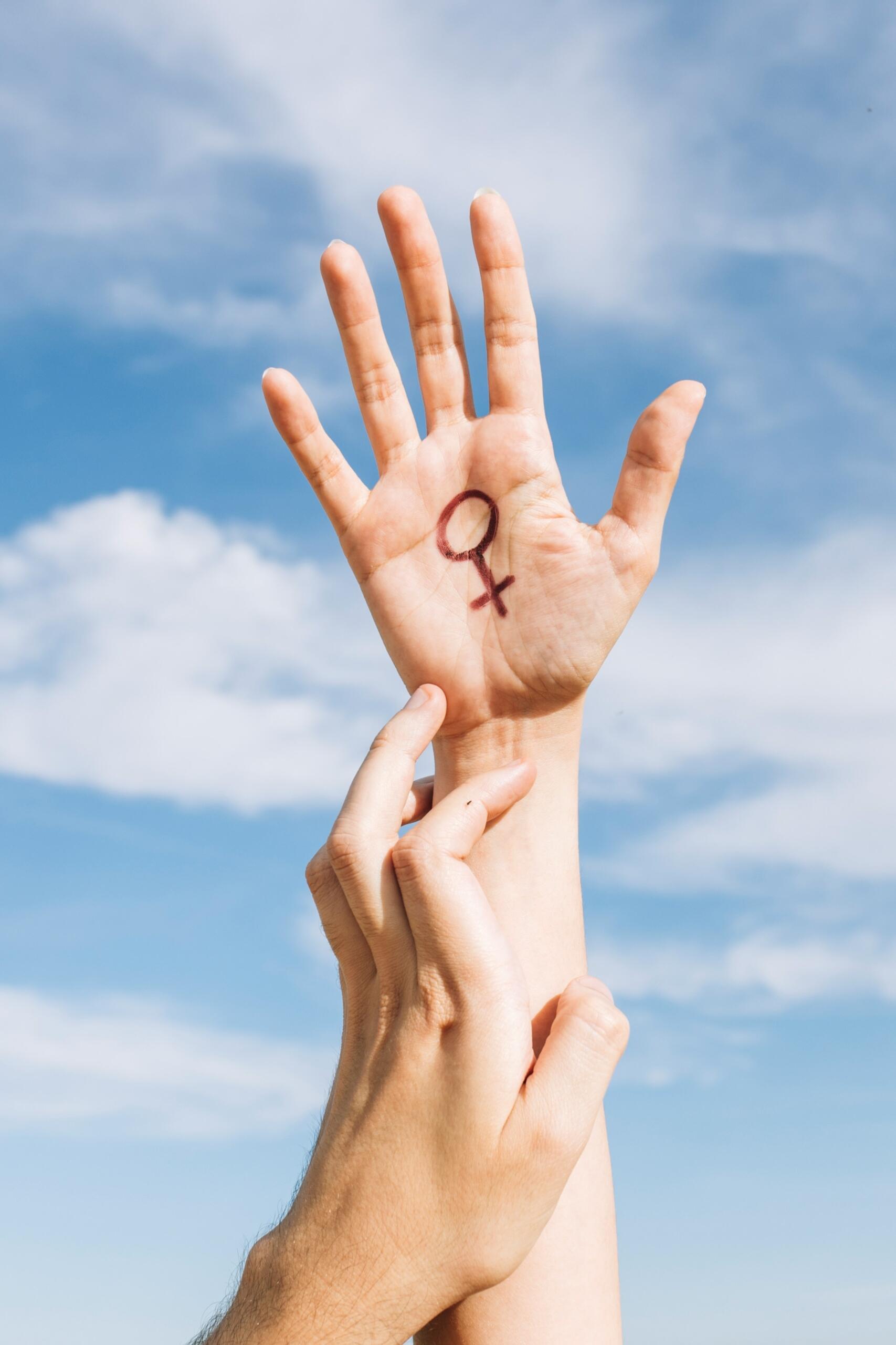 Una mano con un símbolo femenino dibujado en la palma se alza hacia el cielo azul, mientras otra mano la toca suavemente.
