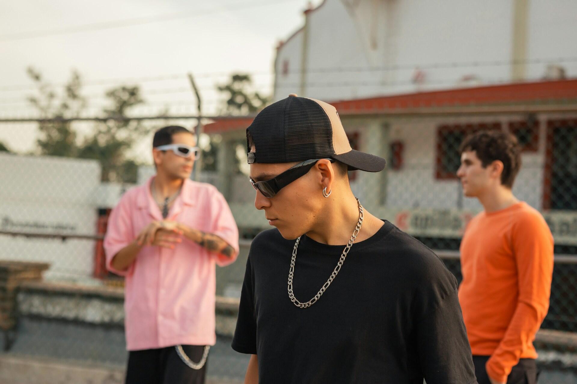 Tres jóvenes están de pie al aire libre con ropa informal: una camiseta negra, una camisa rosa y una camiseta naranja de manga larga, con una cerca y árboles al fondo.