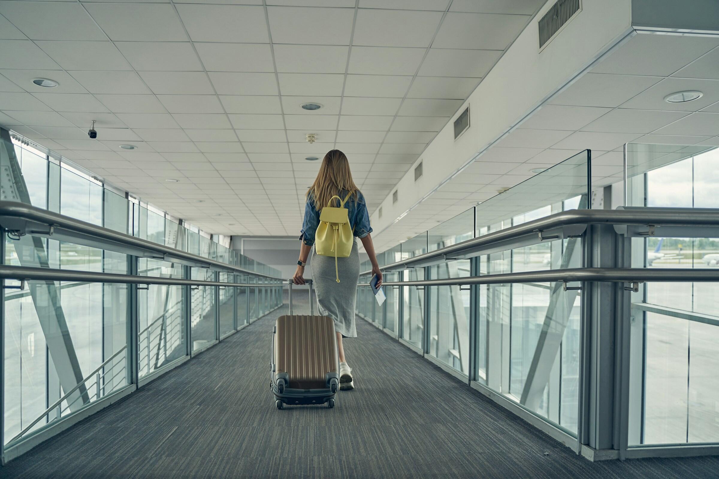 una mujer caminando por un pasillo en lo que parece ser un aeropuerto. Vemos su espalda y lleva una mochila amarilla y una maleta. Tiene un pasaporte en la mano y está vestida con ropa informal. El pasillo tiene grandes ventanas con vistas exteriores.