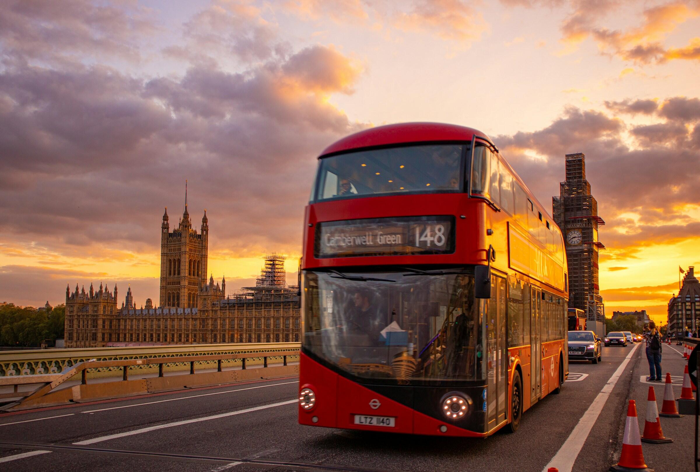 Un autobús rojo de dos pisos circula por una concurrida calle, con el emblemático Big Ben de Londres y una colorida puesta de sol al fondo.