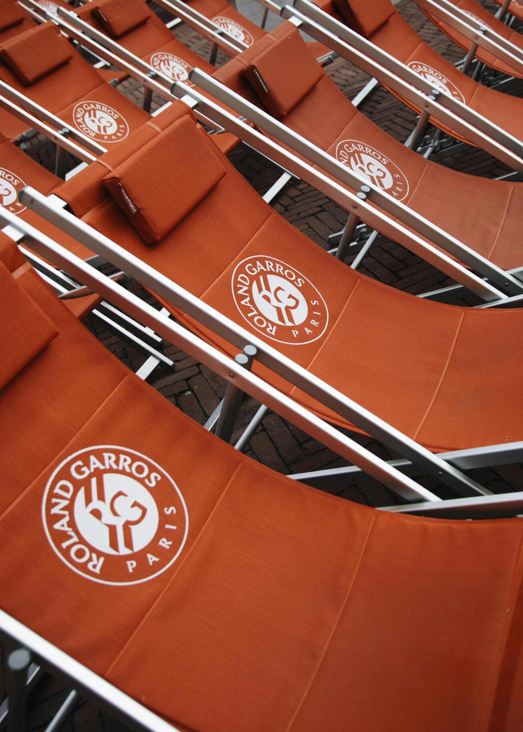 Una fila de tumbonas naranjas con el logotipo «Roland Garros Paris», dispuestas sobre una superficie de ladrillo, invitan al descanso en el evento tenístico.