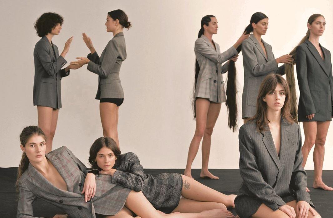 Varias mujeres, vestidas con chaquetas holgadas, posan inexpresivas. Algunas interactúan en parejas, mientras que otras yacen en el suelo, creando una atmósfera tranquila.