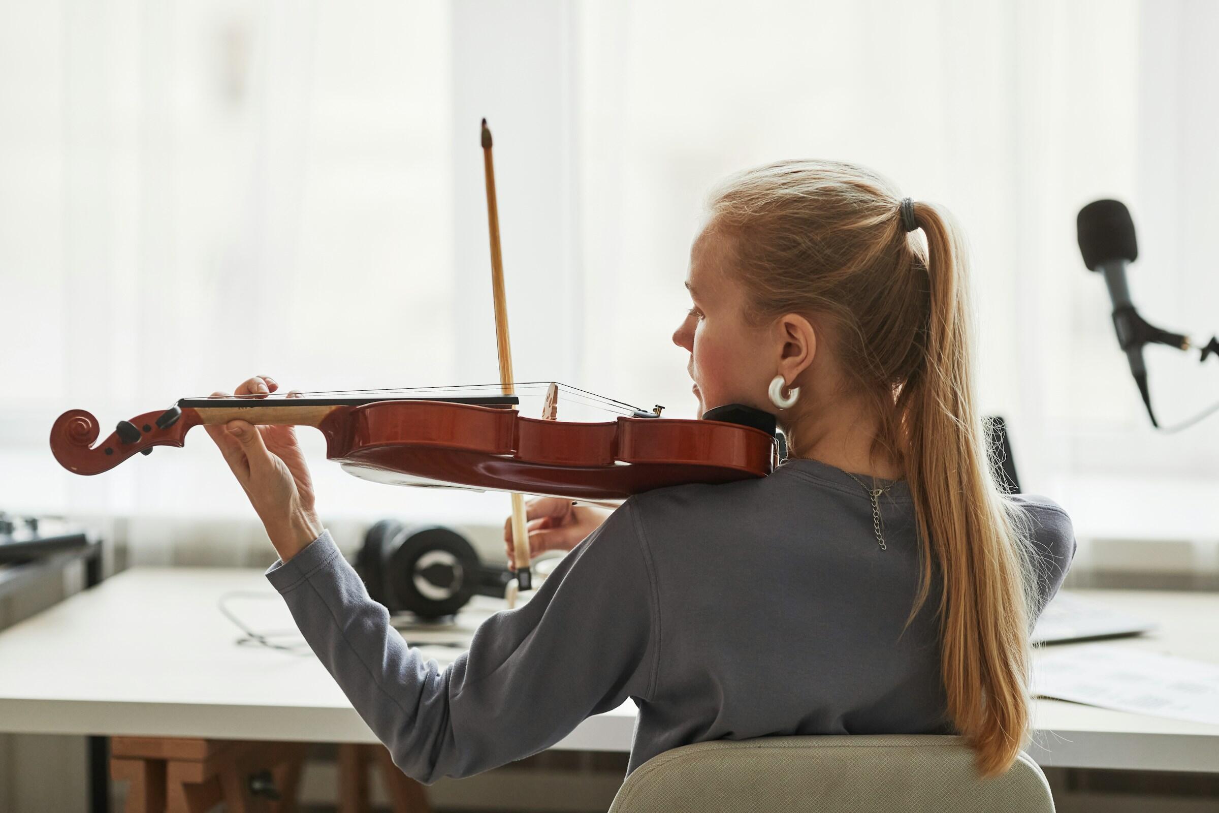 Un joven músico sostiene un violín, listo para tocar, en una sala luminosa y moderna con equipos musicales visibles al fondo.