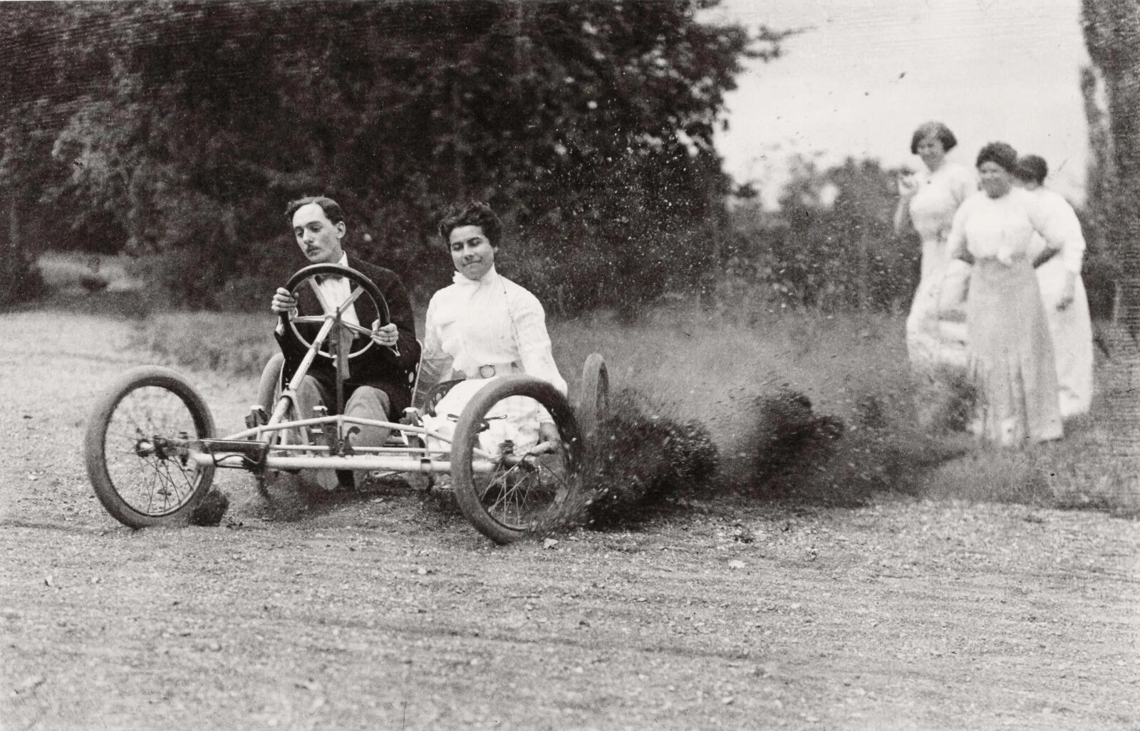 Un coche antiguo con un hombre y una mujer conduce por un camino de tierra, levantando una nube de polvo. Tres mujeres vestidas con trajes de época observan desde un lado, evocando un ambiente divertido y aventurero.
