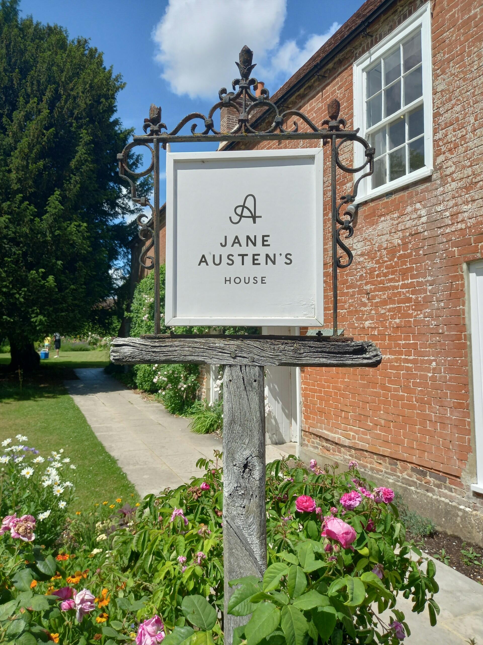 Cartel de la casa de Jane Austen, con una elegante tipografía, enmarcado por vegetación y flores de colores, sobre un cielo azul claro.
