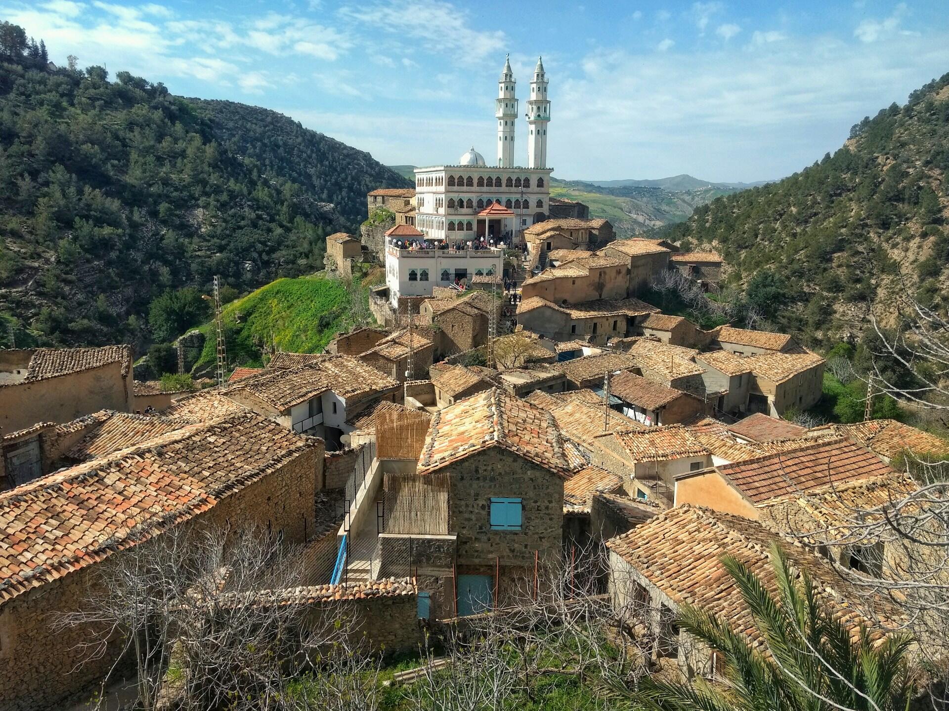 Vista desde la azotea de un pueblo situado en una ladera, con tejados rústicos de terracota y una prominente mezquita con dos minaretes gemelos. Todo ello rodeado de una exuberante vegetación, bajo un cielo azul intenso.